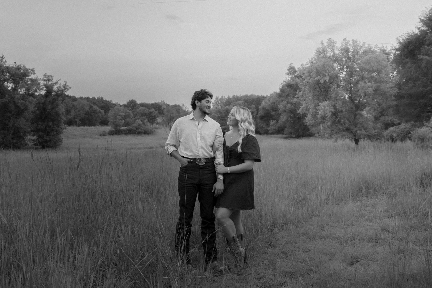 Black and white photo of a couple standing together in a grassy field at dusk, surrounded by trees.