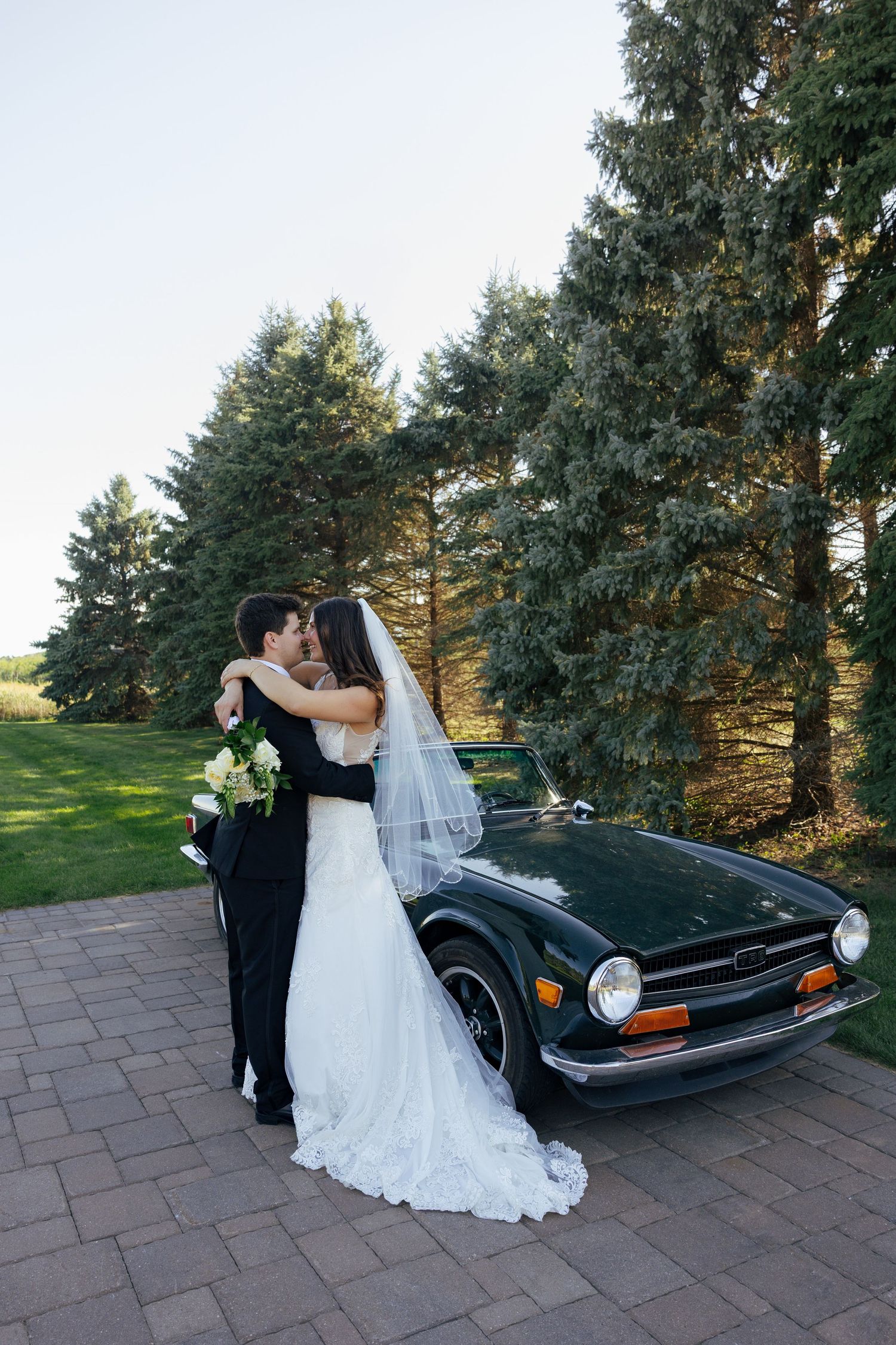 Bride and groom embrace and kiss beside a classic vintage dark green car surrounded by tall evergreen trees on their wedding day.