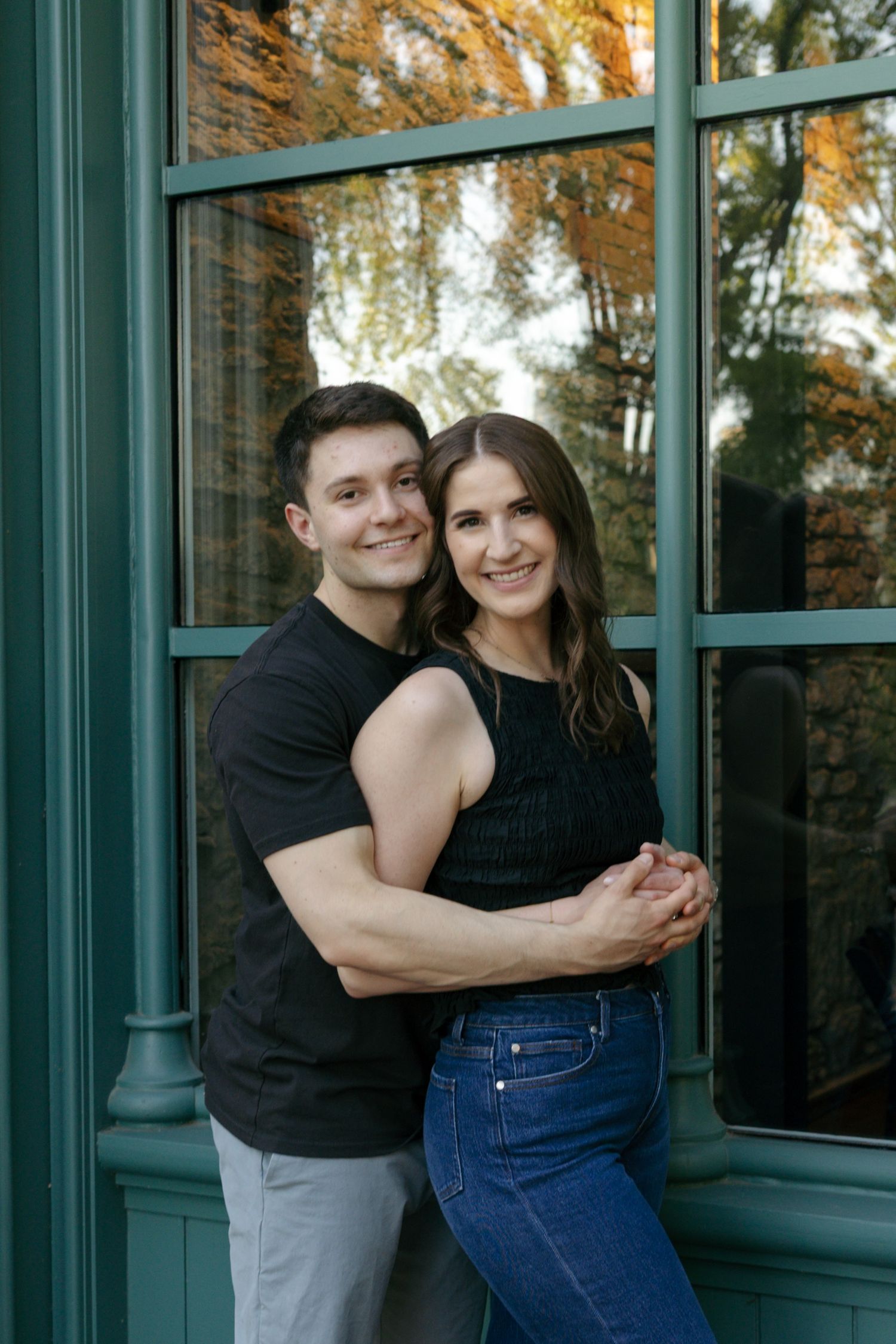 A smiling couple embraces in front of a green-framed window with autumn trees reflected in the glass.