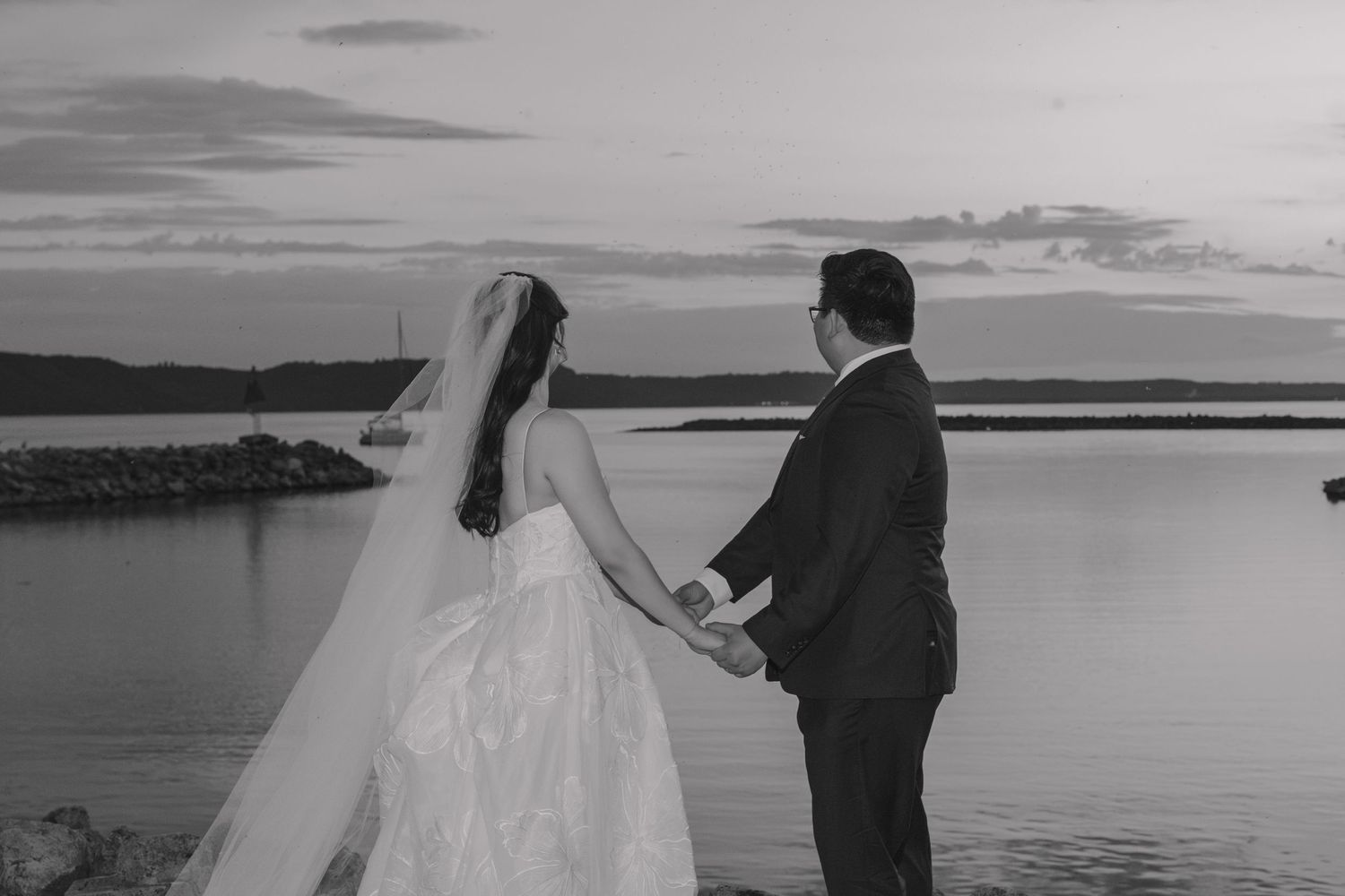 Black and white photo of a bride and groom holding hands by a serene waterfront at sunset.