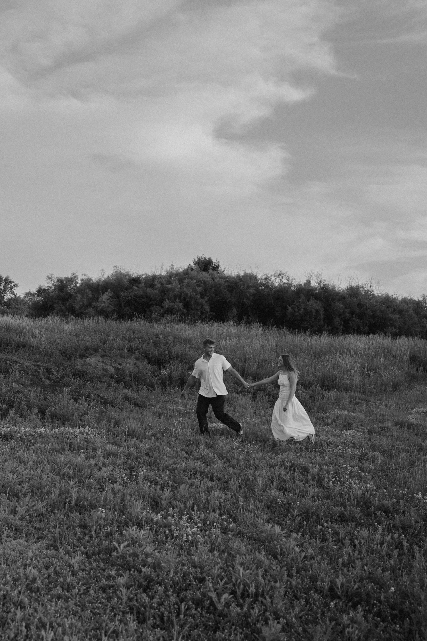 Black and white photo of a couple holding hands while walking through a lush green field under a cloudy sky.