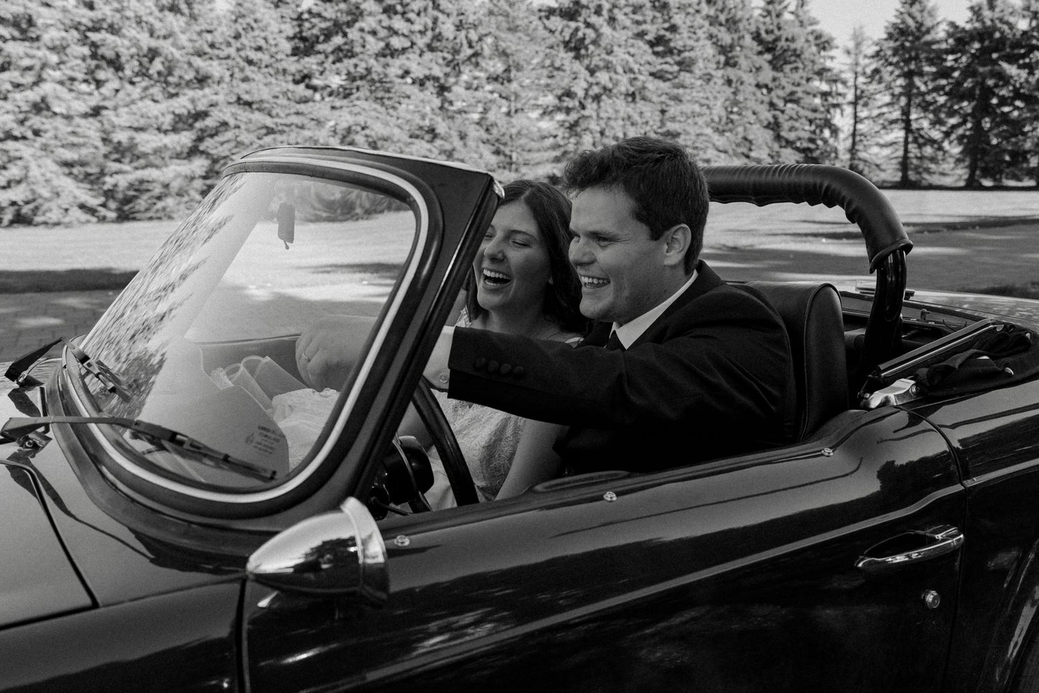 A couple in formal wedding attire smiles joyfully in a classic black convertible car surrounded by snowy trees.