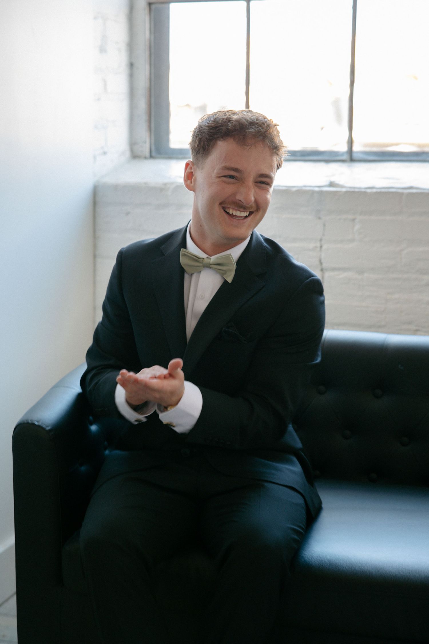 Smiling groom in a black tuxedo with a sage green bow tie sits on a black leather sofa.