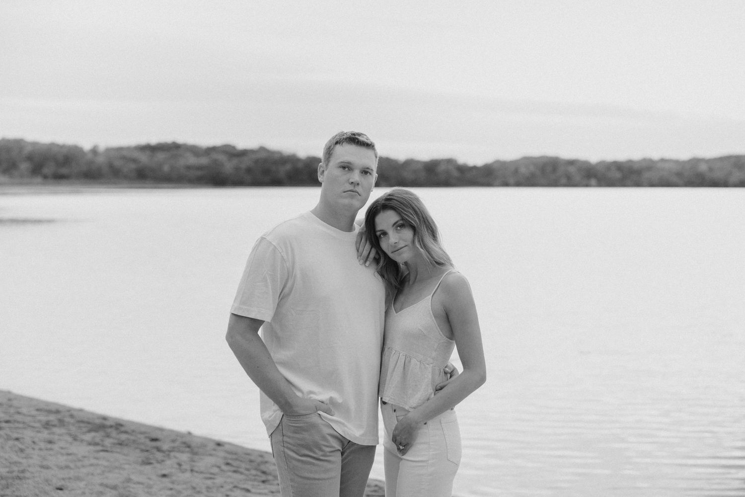 Black and white photo of a couple posing by a lake on a sandy beach with trees in the background.