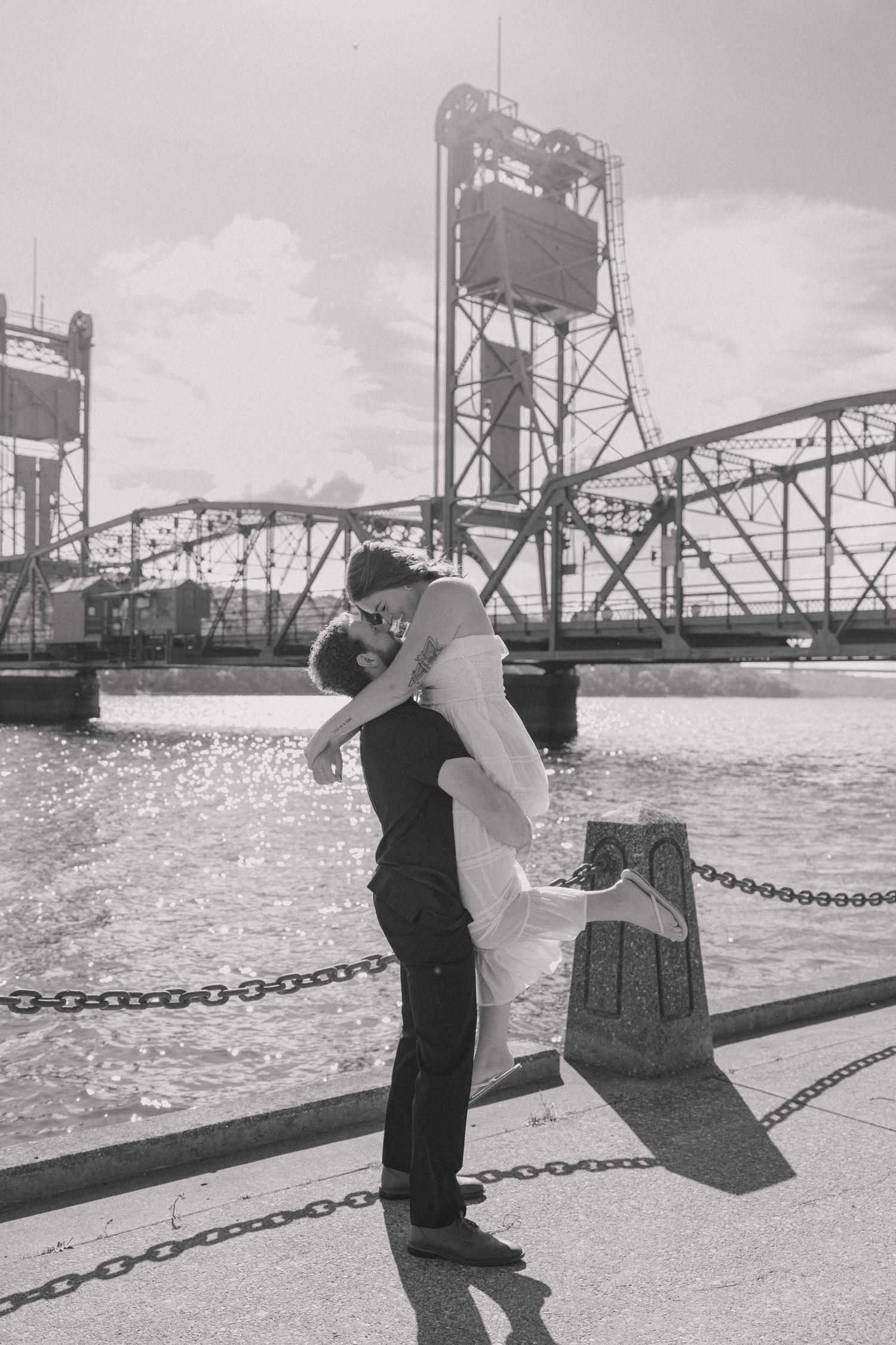 Black and white photo of groom lifting bride for a kiss on a waterfront dock with a steel lift bridge in the background.