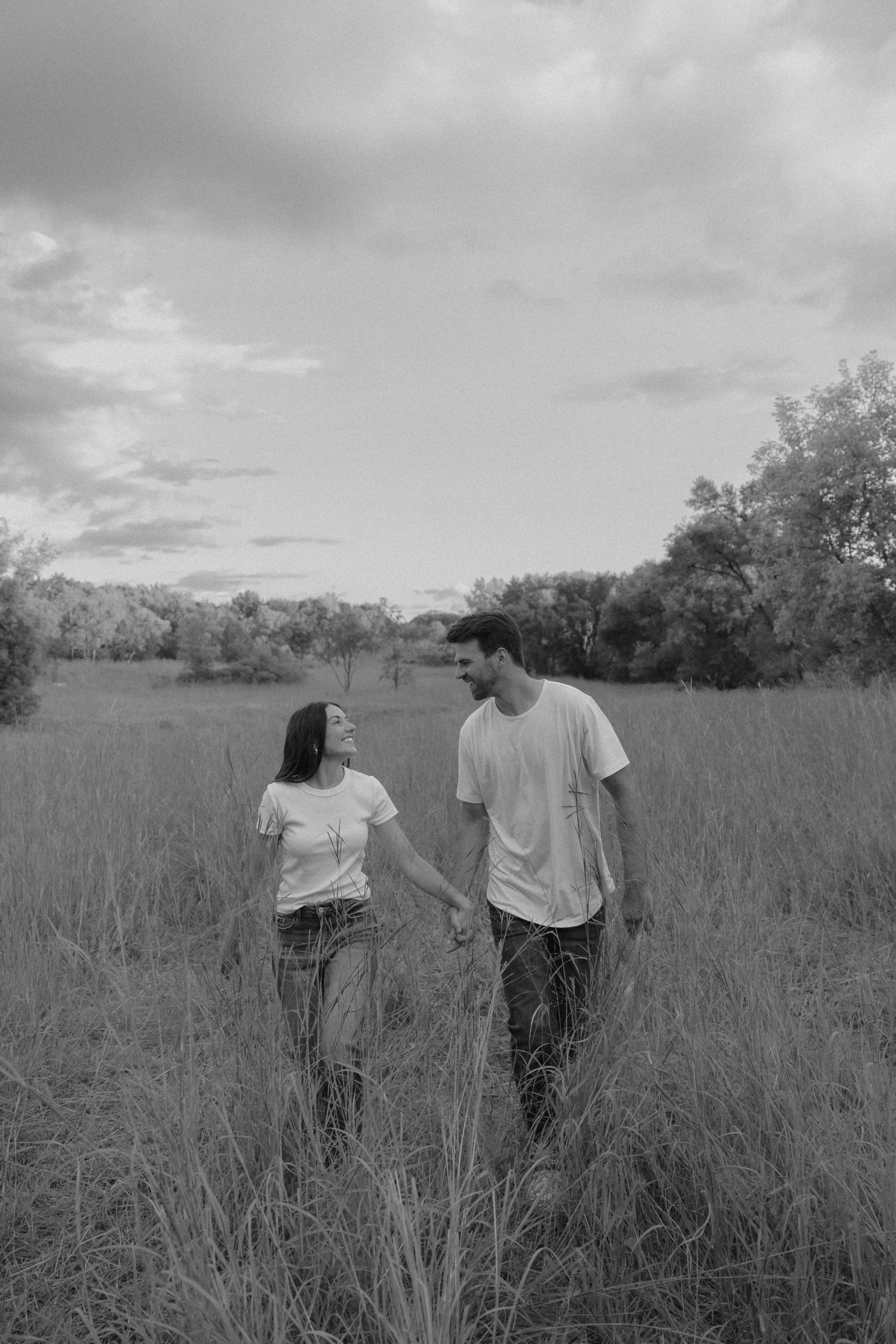 Black and white photo of a couple holding hands walking through a grassy field with trees in the background.