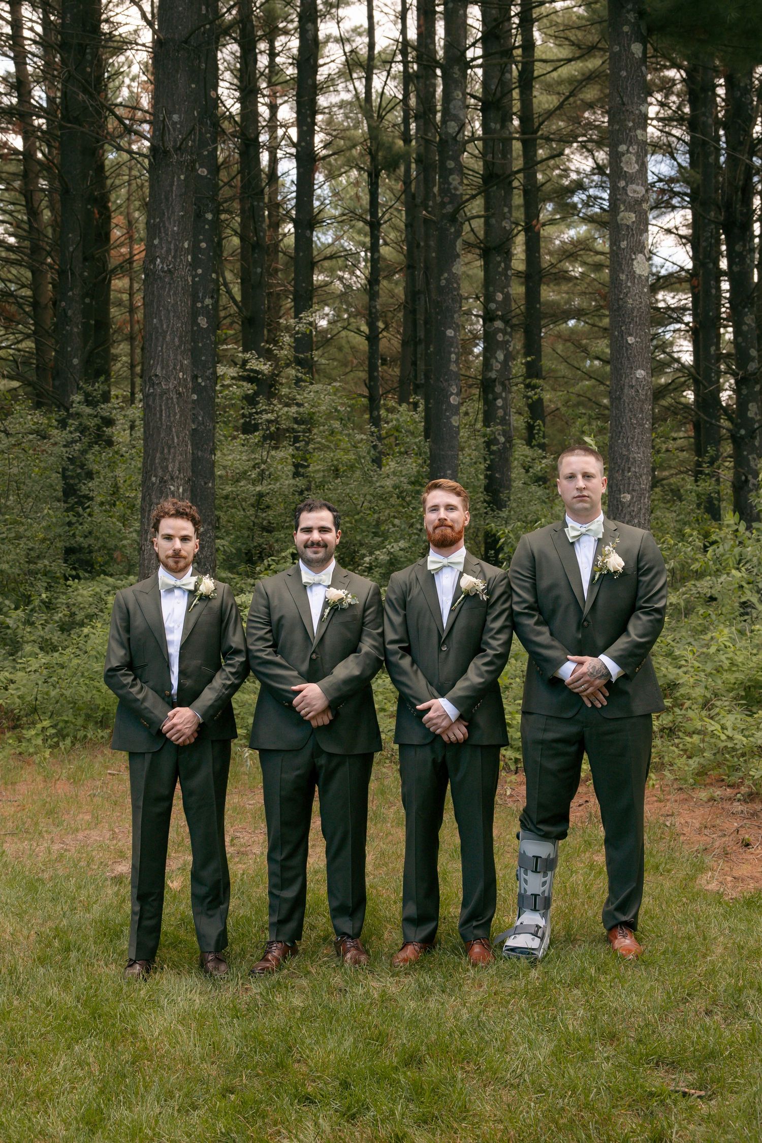Four groomsmen in dark olive suits with boutonnieres pose outdoors in a forest setting, one wearing a medical boot.