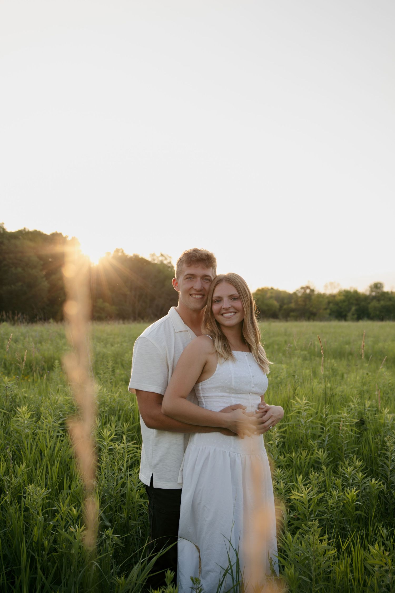 Couple embracing in a sunlit green field at golden hour, woman in white dress, man in white shirt, smiling outdoors.