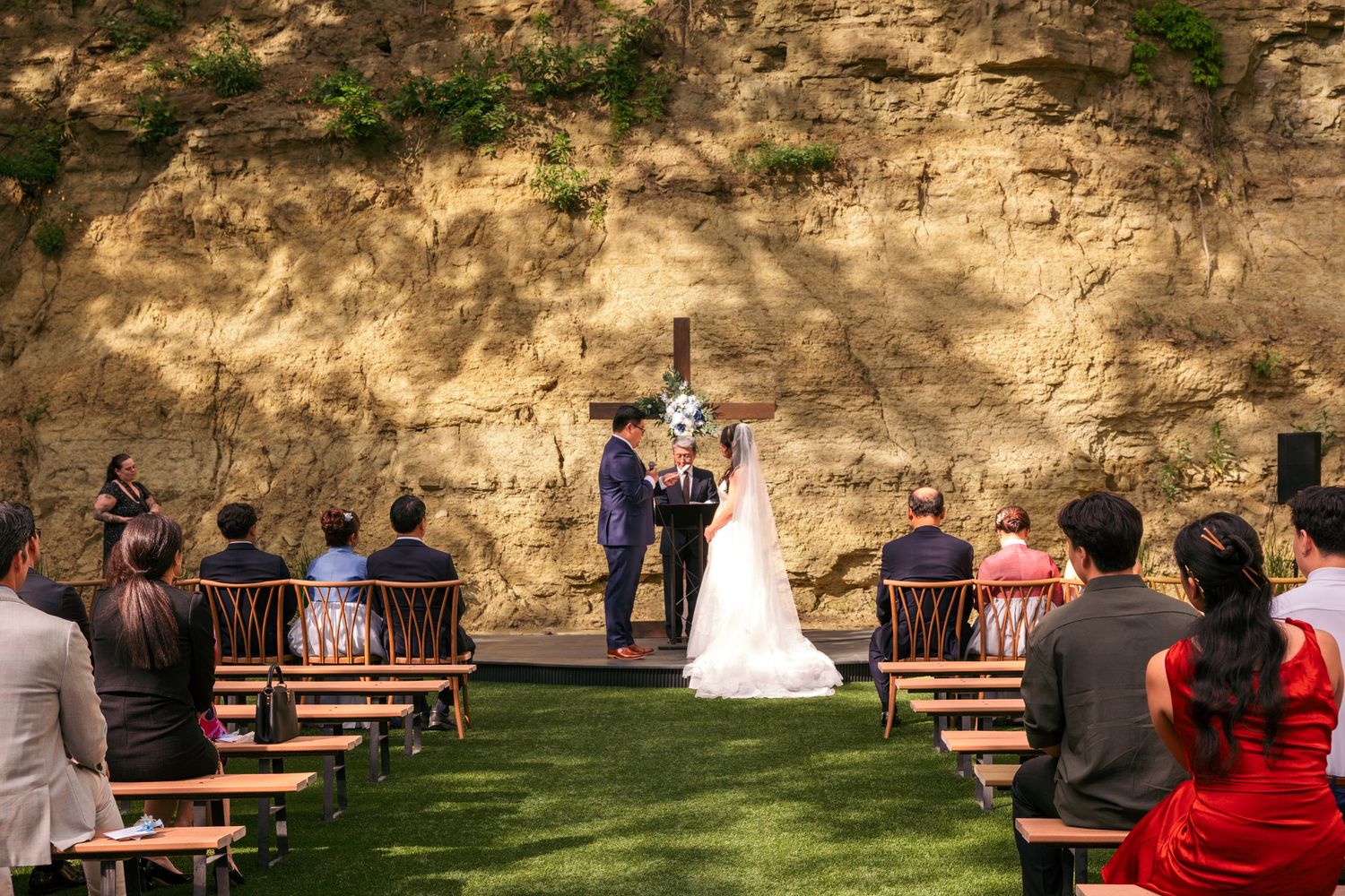 Outdoor wedding ceremony with couple at altar before rustic stone cliff, guests seated on wooden chairs on green lawn.