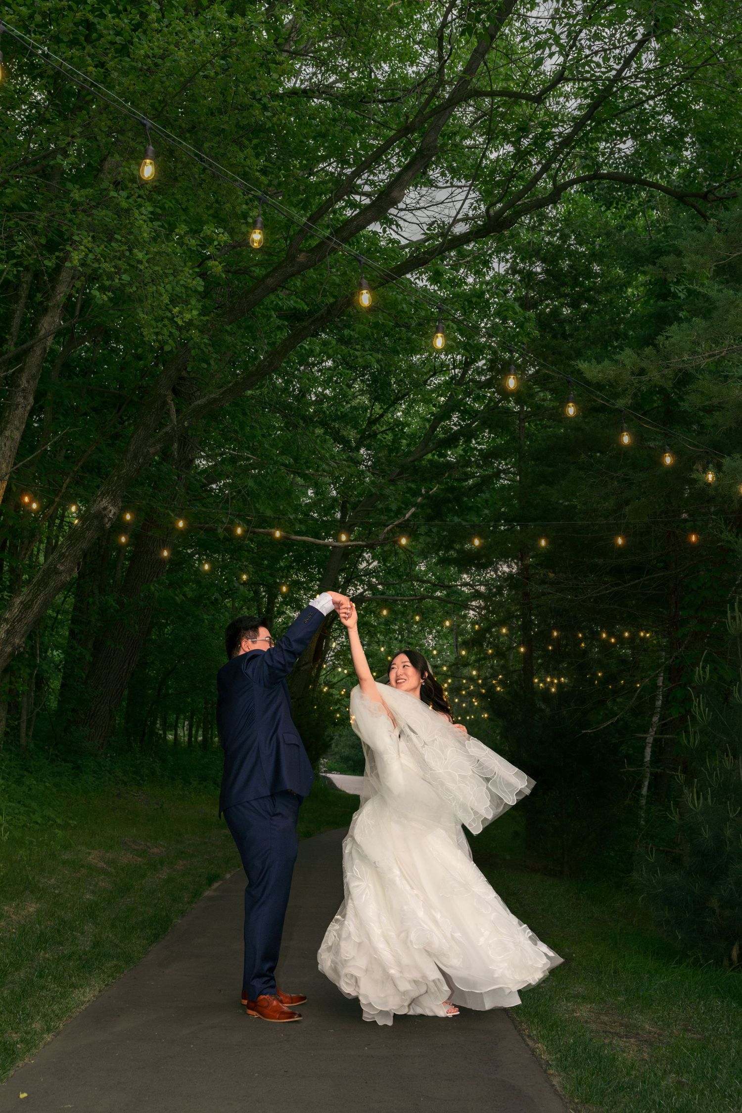 Bride and groom dancing on a tree-lined path adorned with string lights at dusk during outdoor wedding.