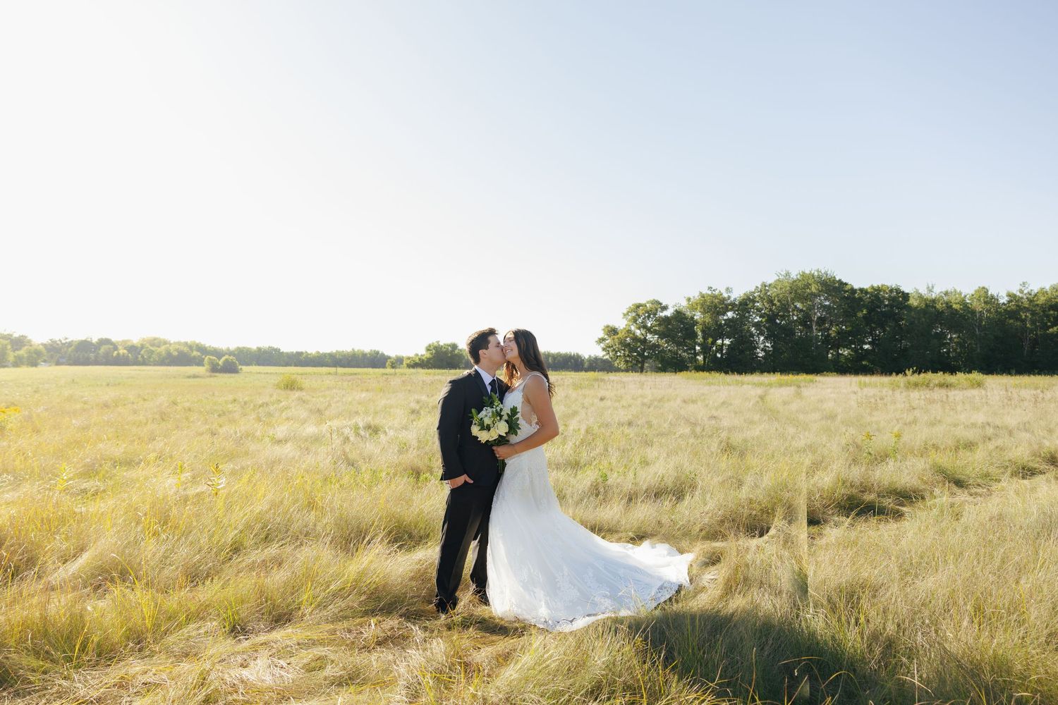 Bride and groom share a kiss in a sunlit golden field with trees in the background on their wedding day.