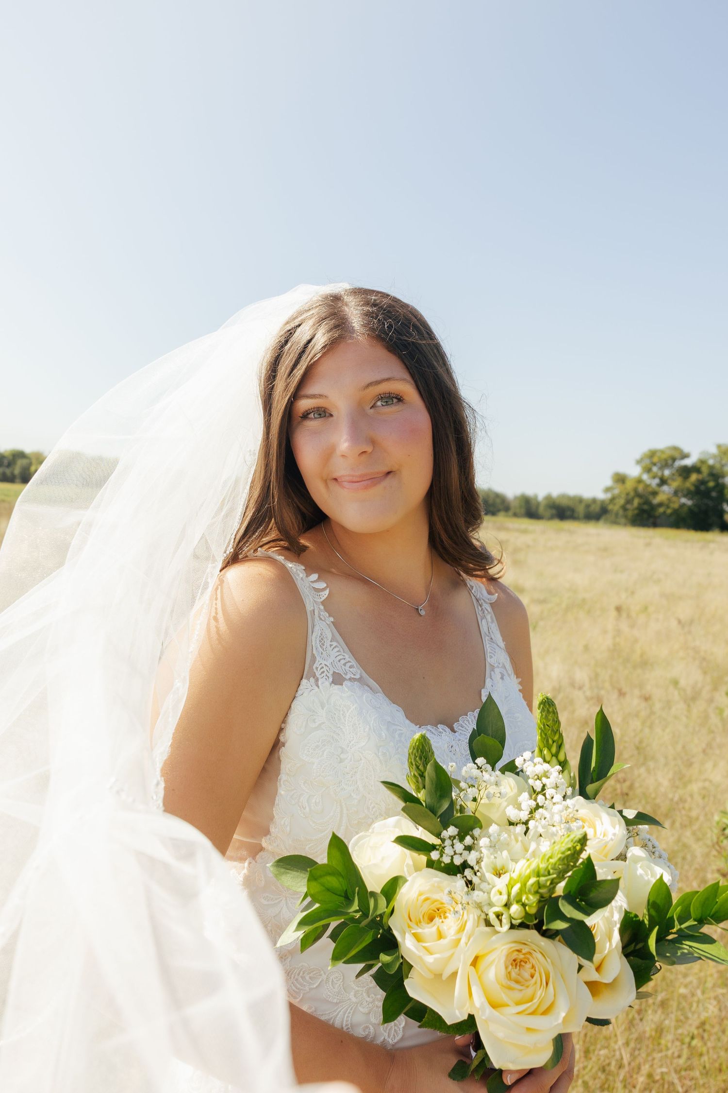 Smiling bride in white lace dress and veil holding bouquet of white roses in sunny field.