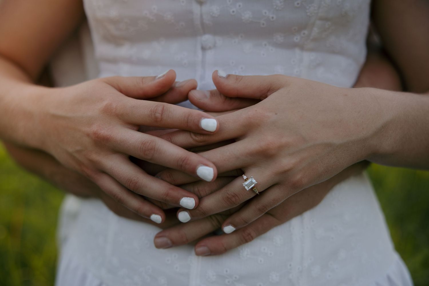 Close-up of two people's hands interlaced, one wearing a diamond engagement ring, with white manicured nails on a white dress.