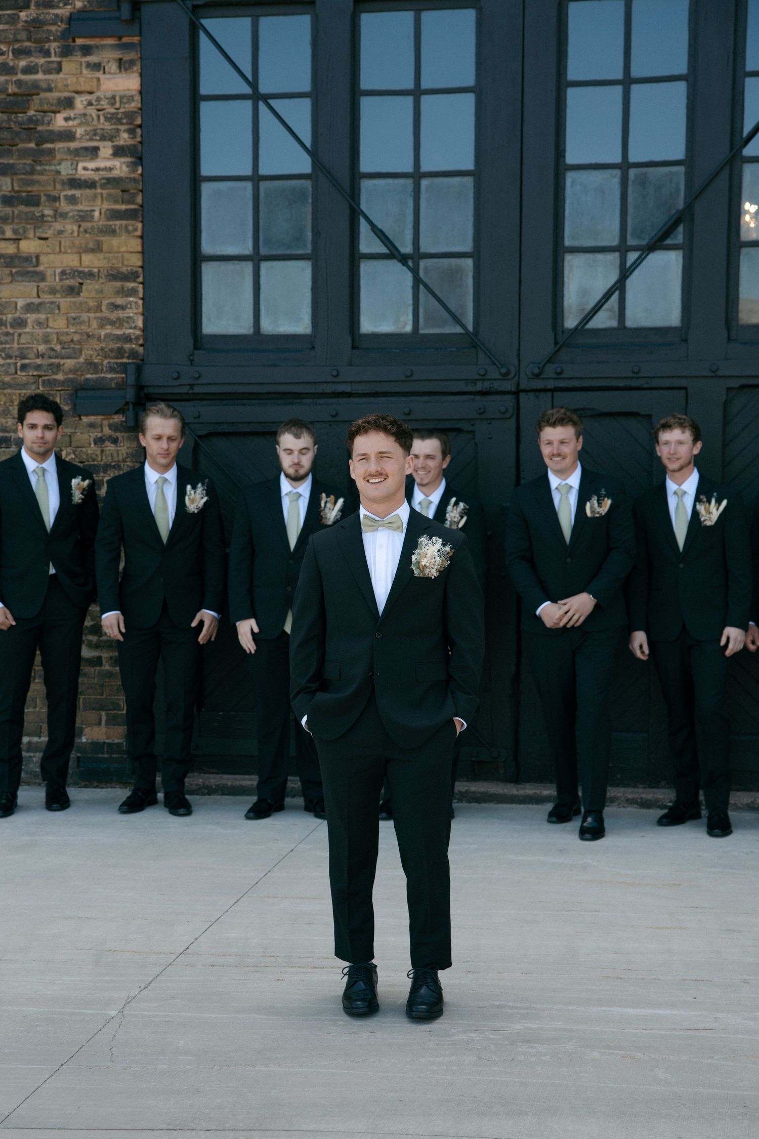 Groom stands confidently in front of groomsmen in black suits outside elegant dark wooden doors at wedding.