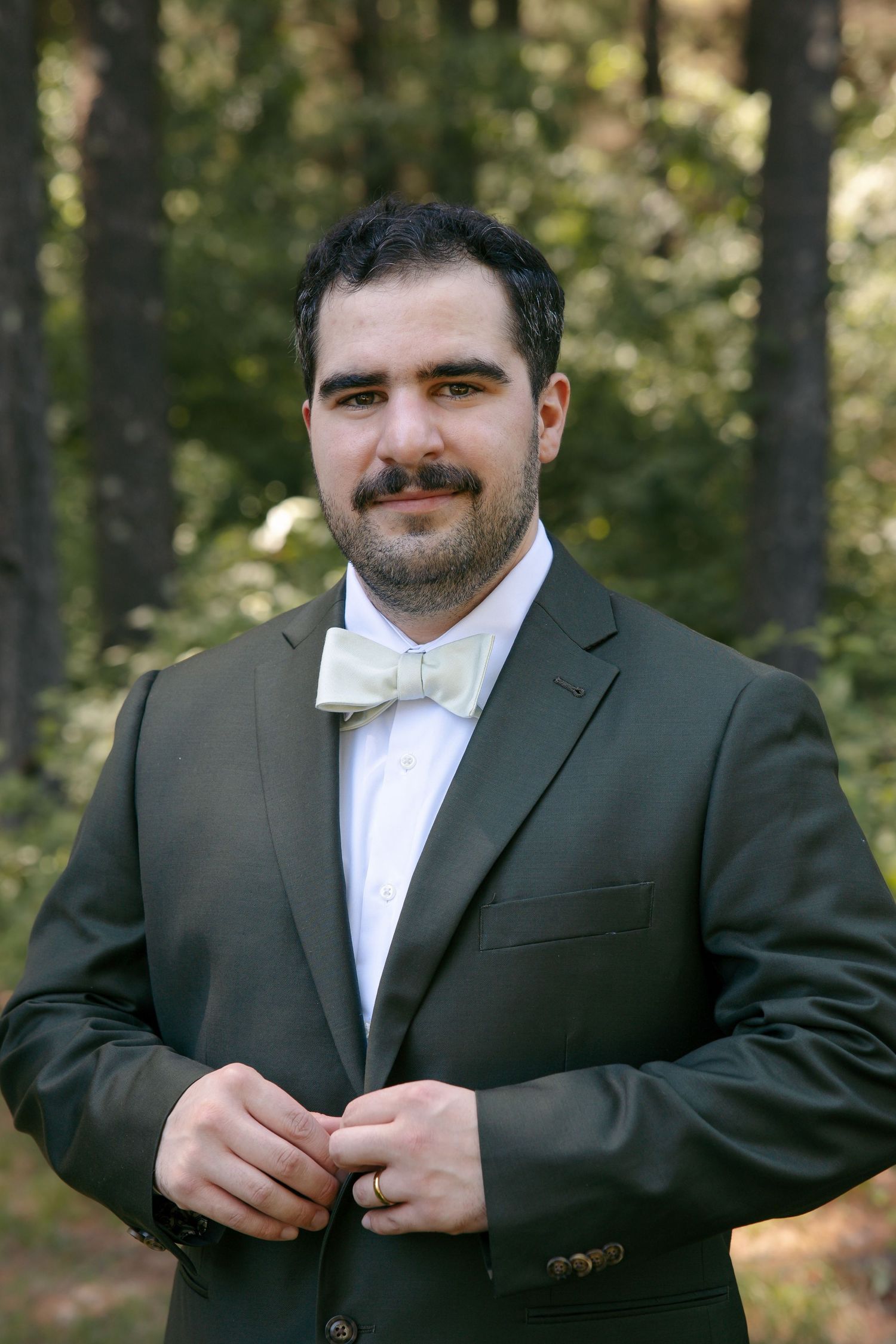 A well-dressed man in a dark green suit and bow tie stands outdoors among trees, adjusting his jacket buttons.