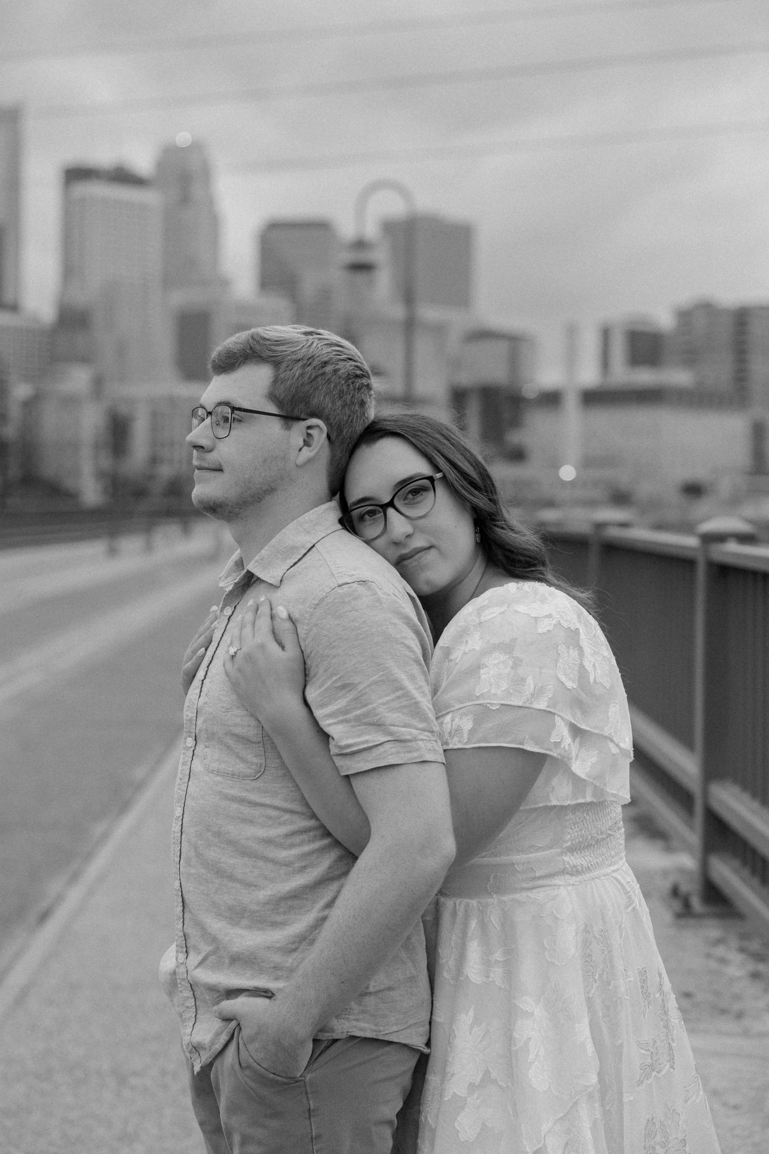 Black and white photo of a couple embracing on a bridge with Minneapolis skyline in the background.