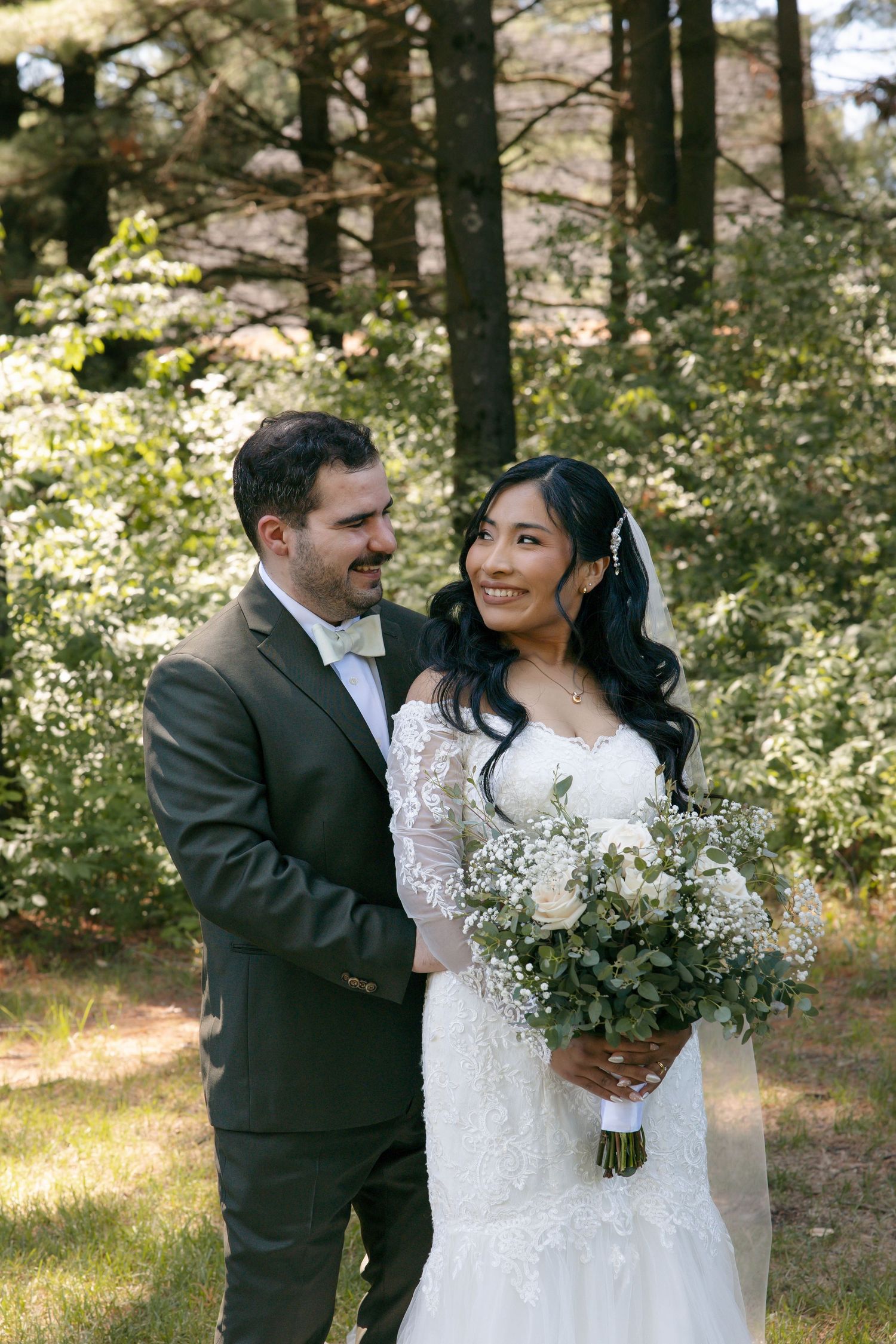 A bride and groom pose together outdoors in a lush green forest, she holds a white floral bouquet in a lace gown.