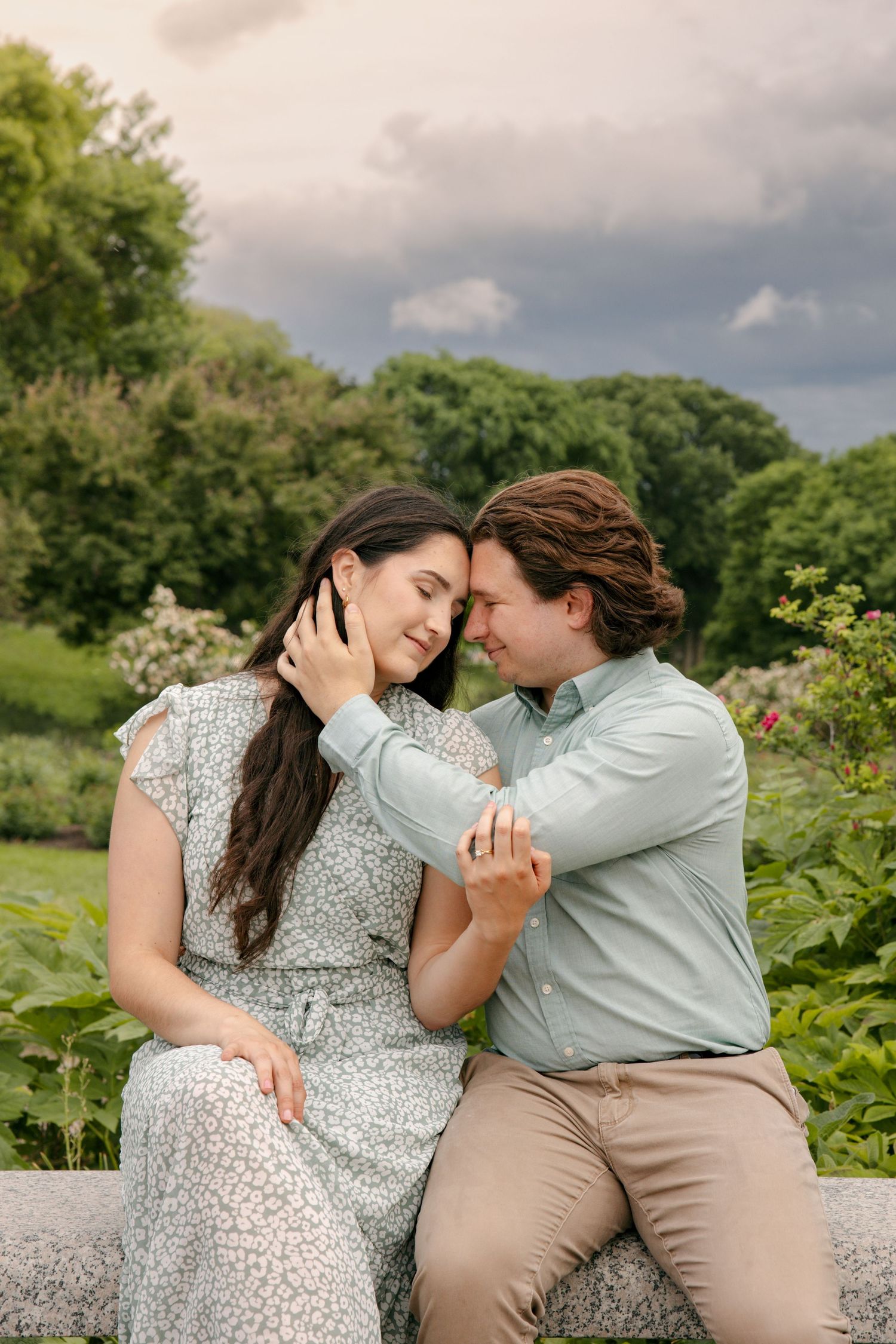 Romantic couple sitting on stone bench in lush garden, man tenderly holding woman's face as they lean foreheads together.