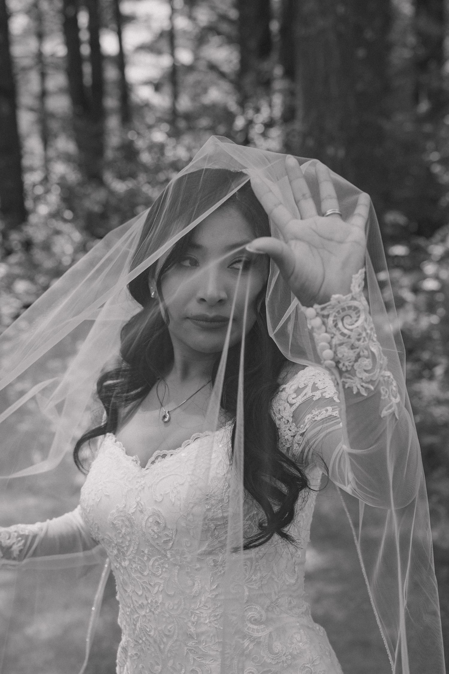 Black and white portrait of a bride lifting her veil in a forest, wearing a lace wedding dress and necklace.