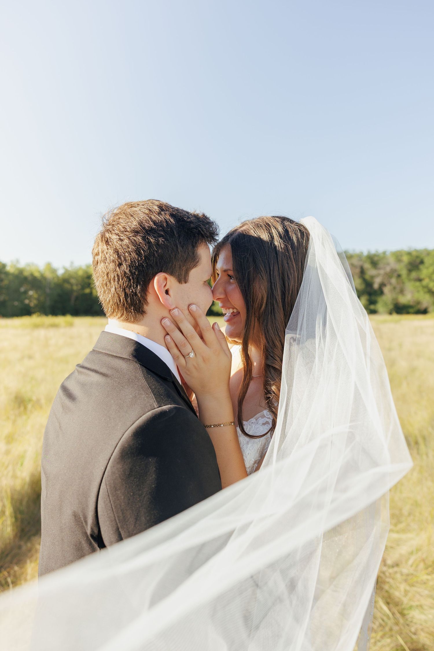 Bride and groom almost kissing in sunny field, wedding veil flowing around them.