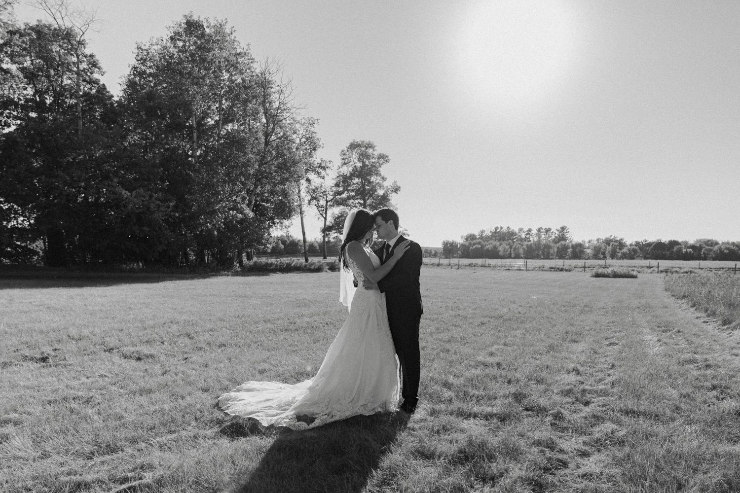 Black and white photo of bride and groom embracing in sunny open field with flowing wedding gown.