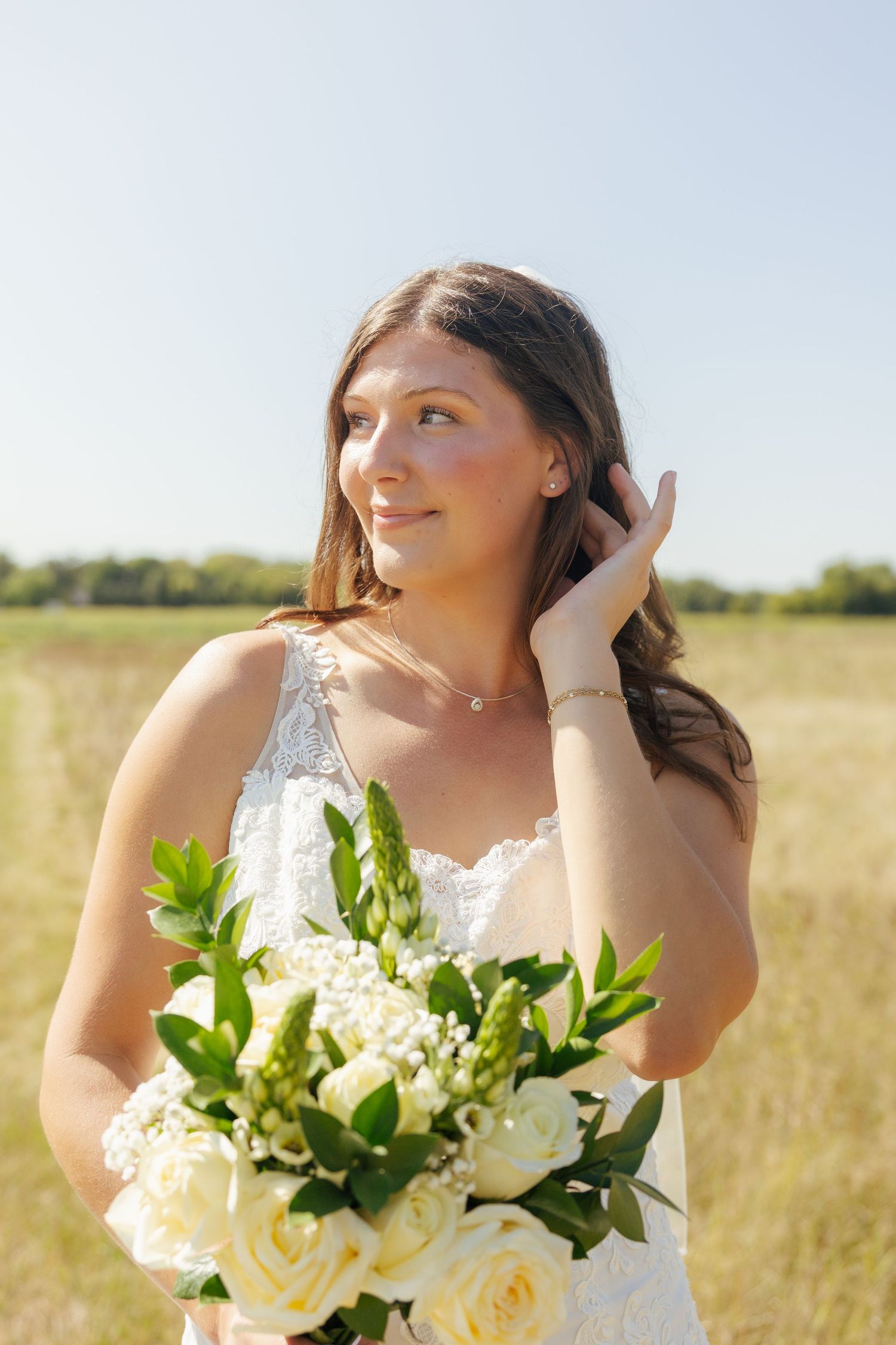 Young bride in white lace dress holding white rose bouquet in sunny field, smiling and looking away.
