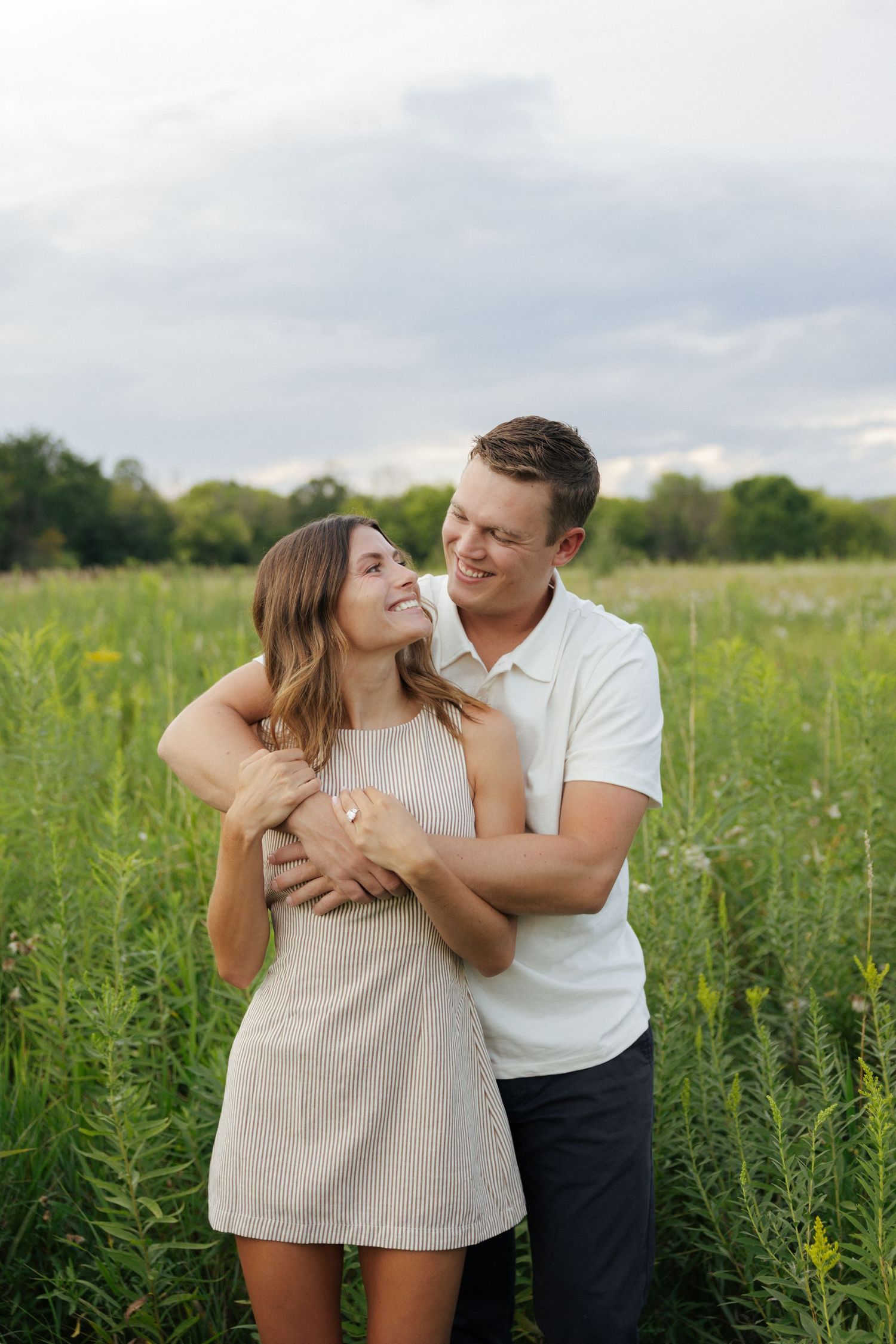 Couple embracing in a lush green field, woman in beige dress smiling up at man in white polo shirt.