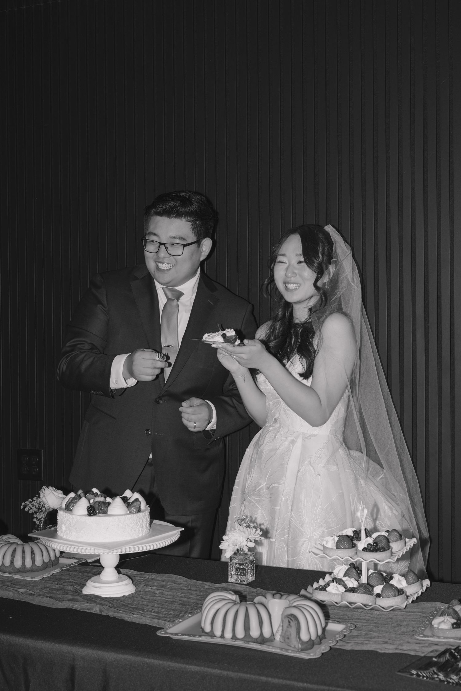 Asian bride and groom smiling while cutting wedding cake at reception table in black and white photo.