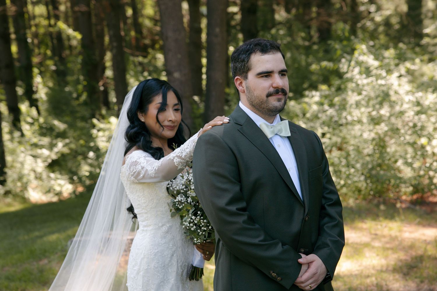 Bride touches grooms shoulder during first look moment in sunlit pine forest wedding.