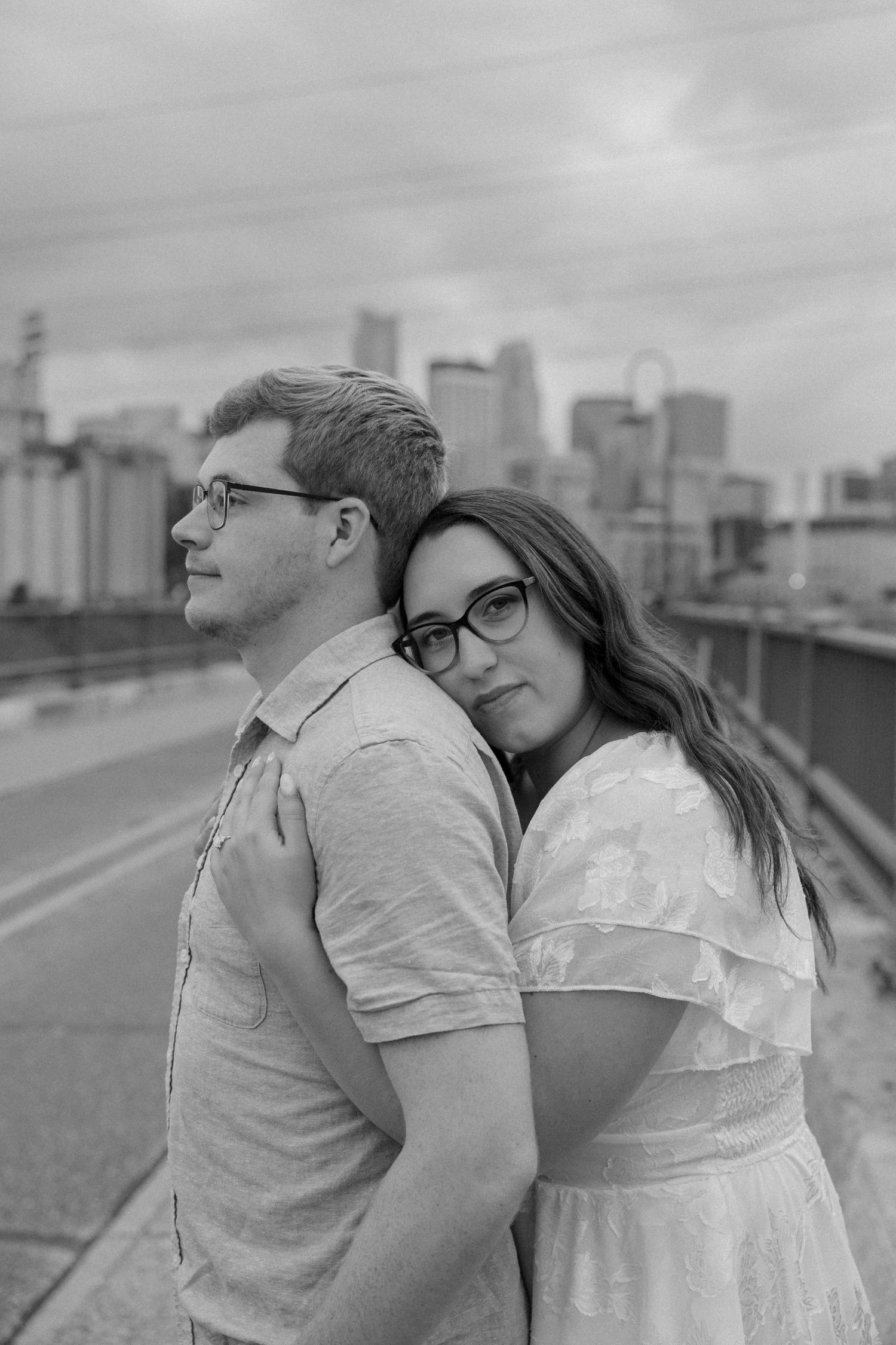 Black and white photo of a couple embracing on a bridge with a city skyline backdrop, both wearing glasses.