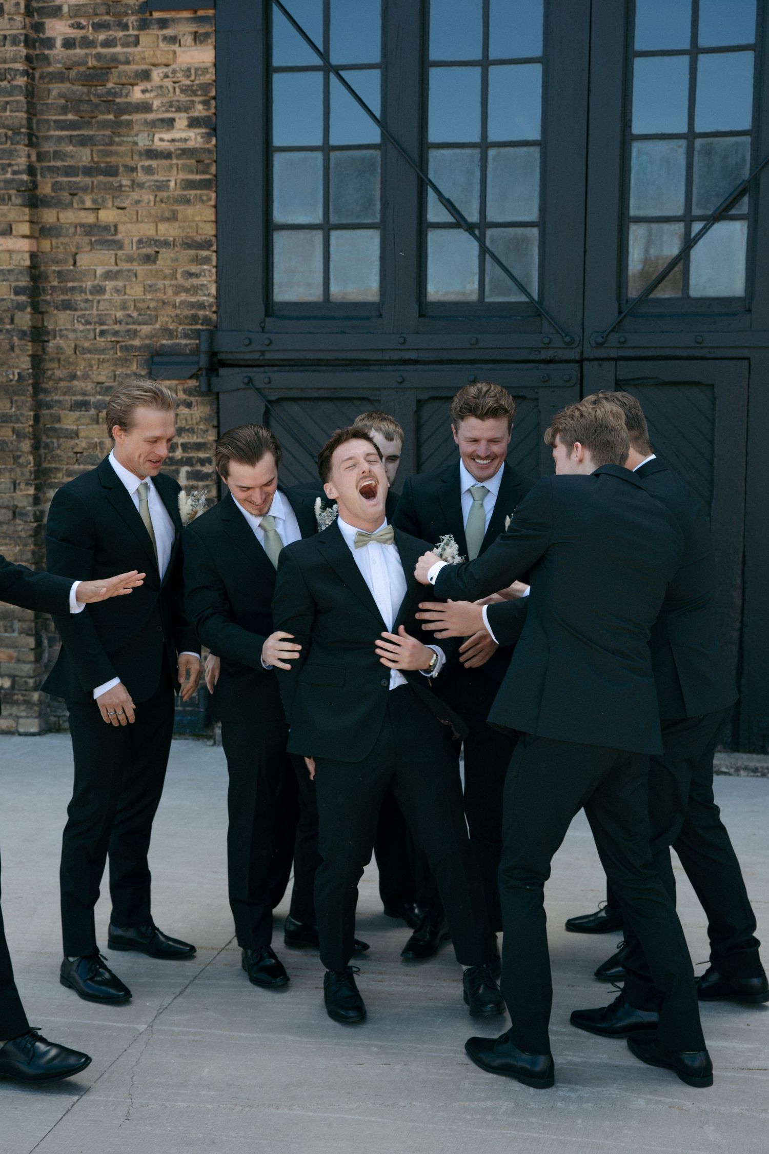 Groom laughing with groomsmen in matching dark suits outside rustic brick venue.