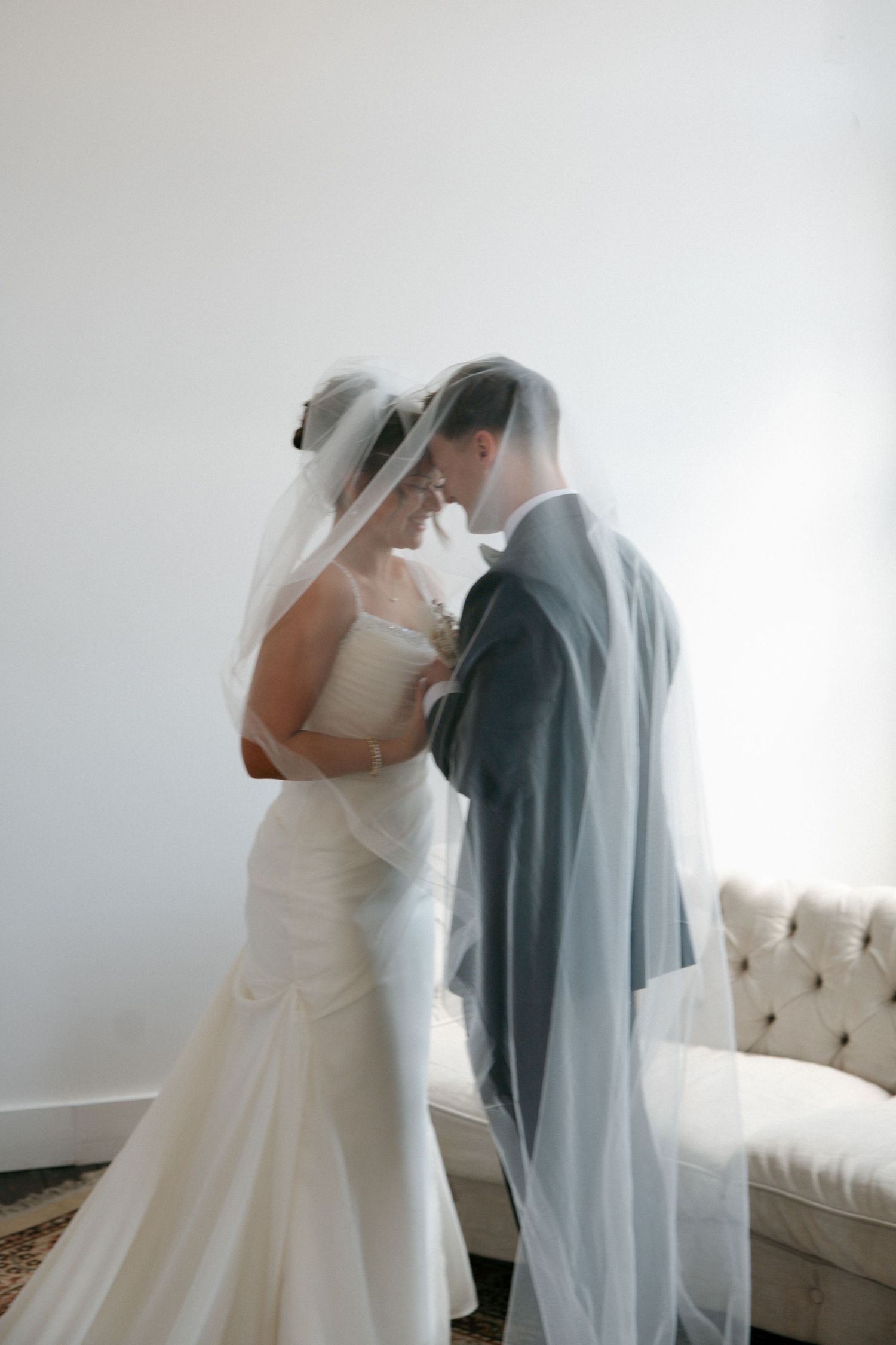 Bride and groom share an intimate moment under a flowing wedding veil in a softly lit room.