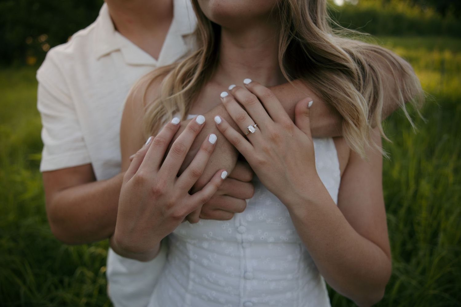 Couple embracing outdoors, woman wearing engagement ring, hands placed over heart in romantic pose.