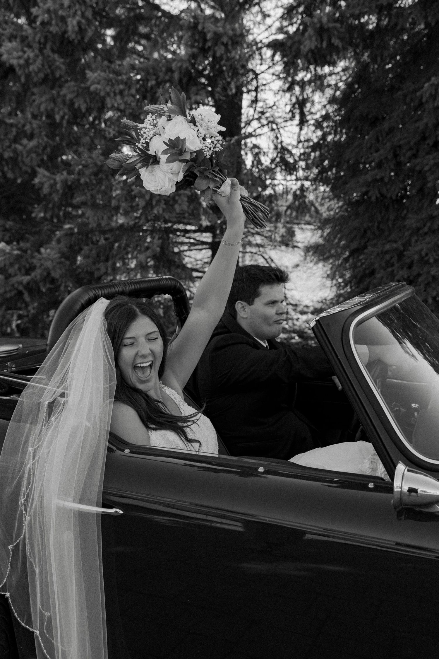 Joyful bride and groom celebrating in a classic convertible car, bride raising bouquet triumphantly in black and white photo.