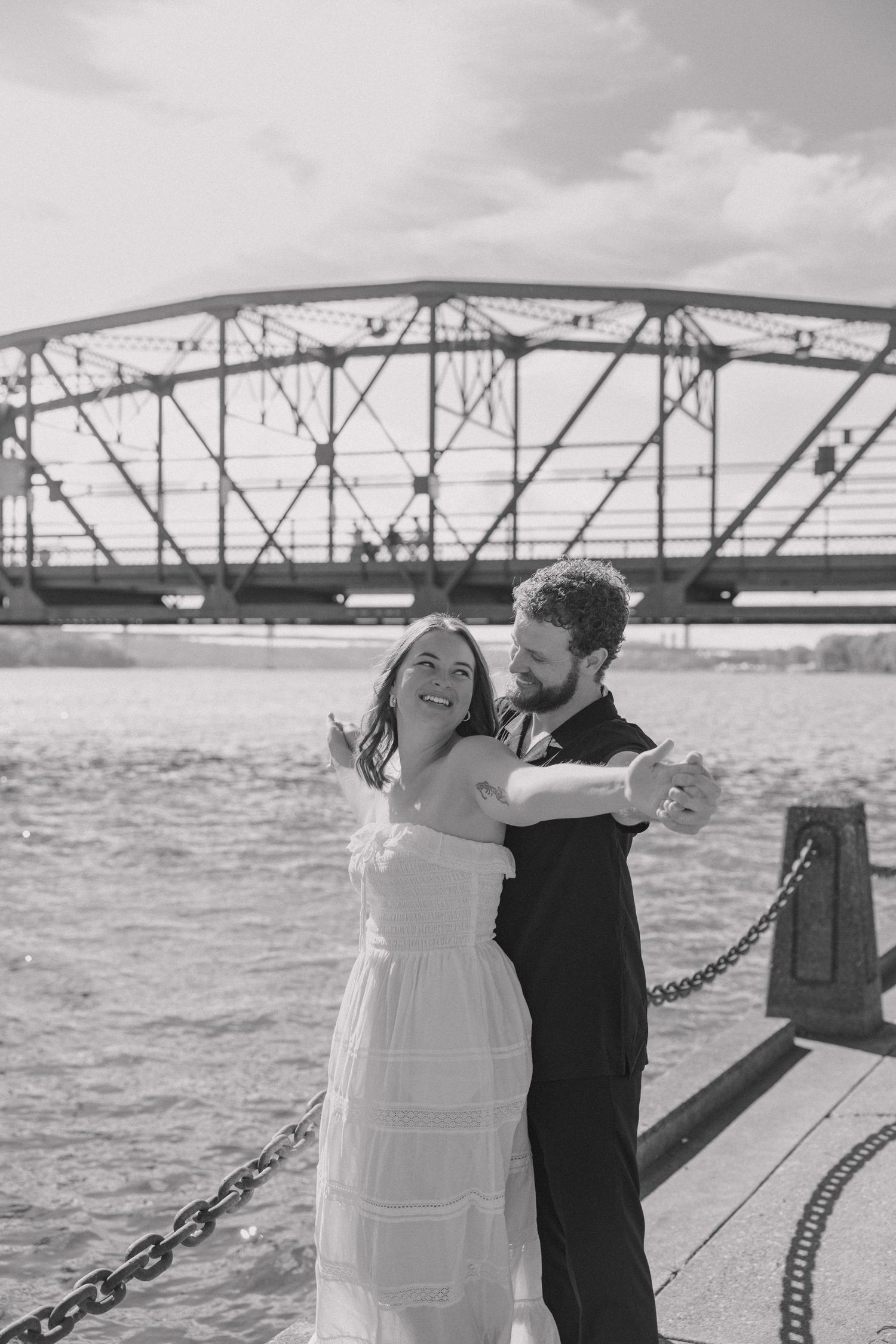 Couple dancing joyfully on a waterfront dock with a steel bridge backdrop in black and white.