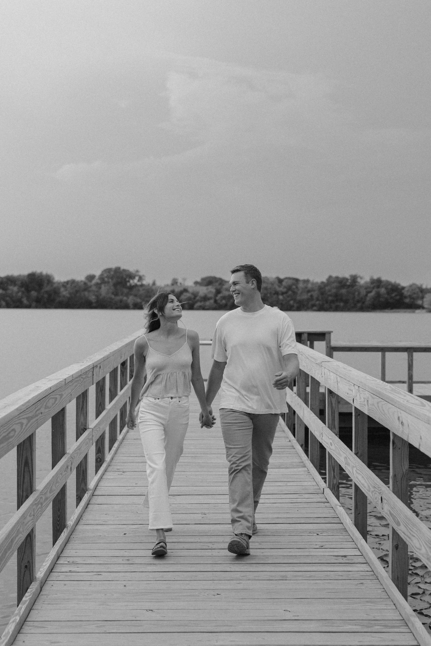 Black and white photo of a couple holding hands and smiling while walking on a wooden pier by a lake.