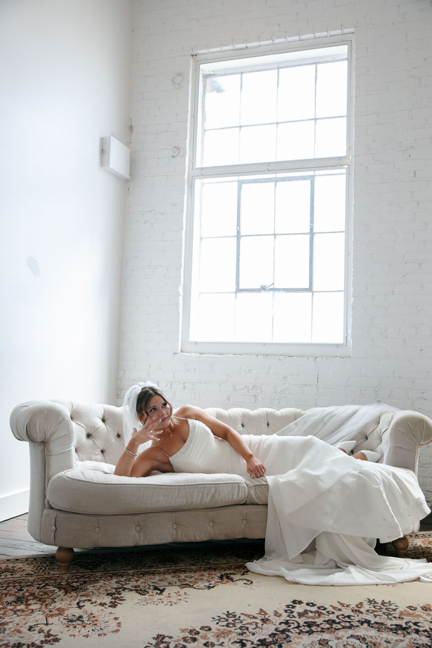 Bride in white wedding dress and veil relaxing on tufted sofa near bright window.