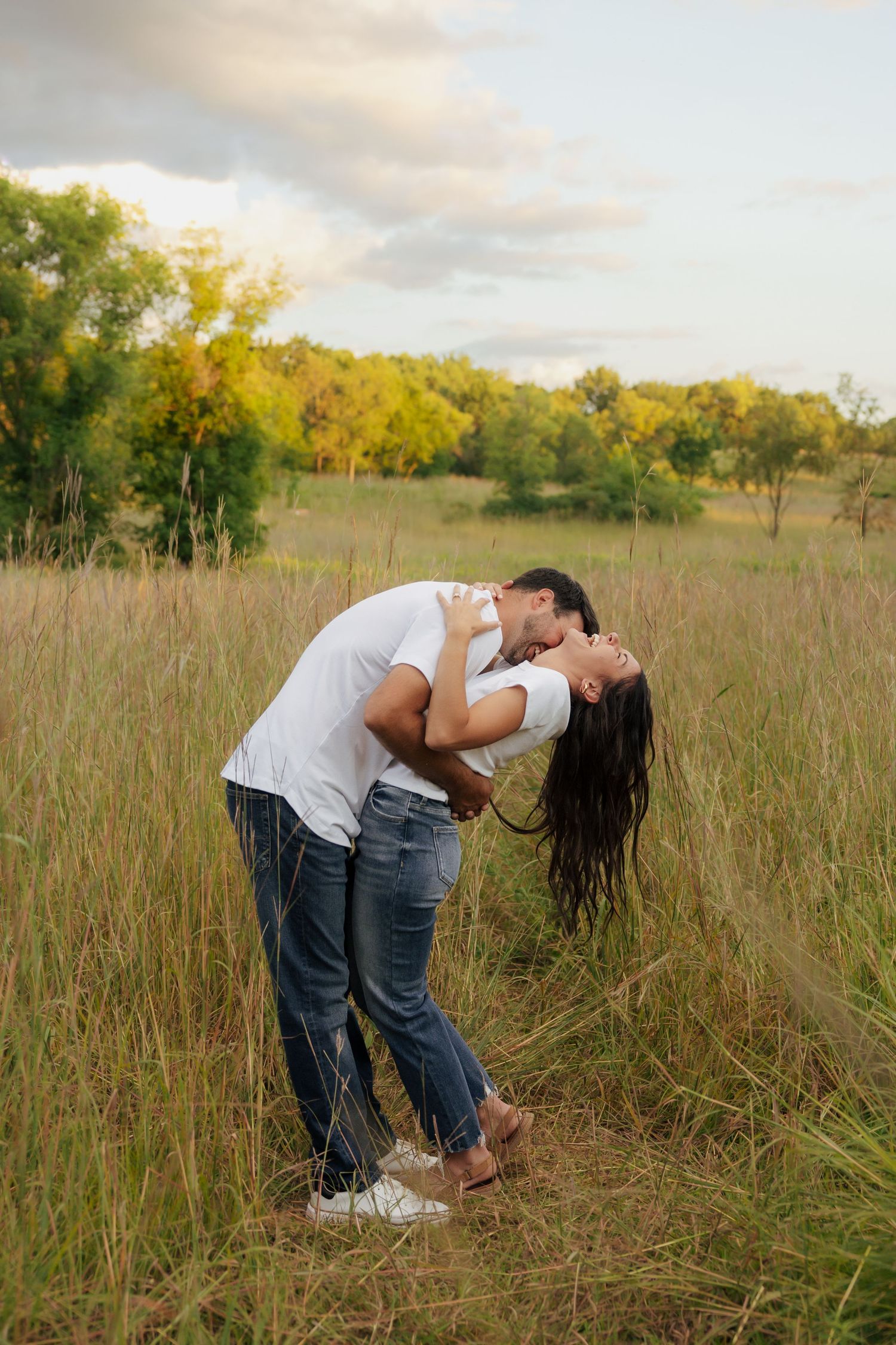 Couple kissing romantically in a grassy field, both wearing white shirts and jeans, with trees in the background.