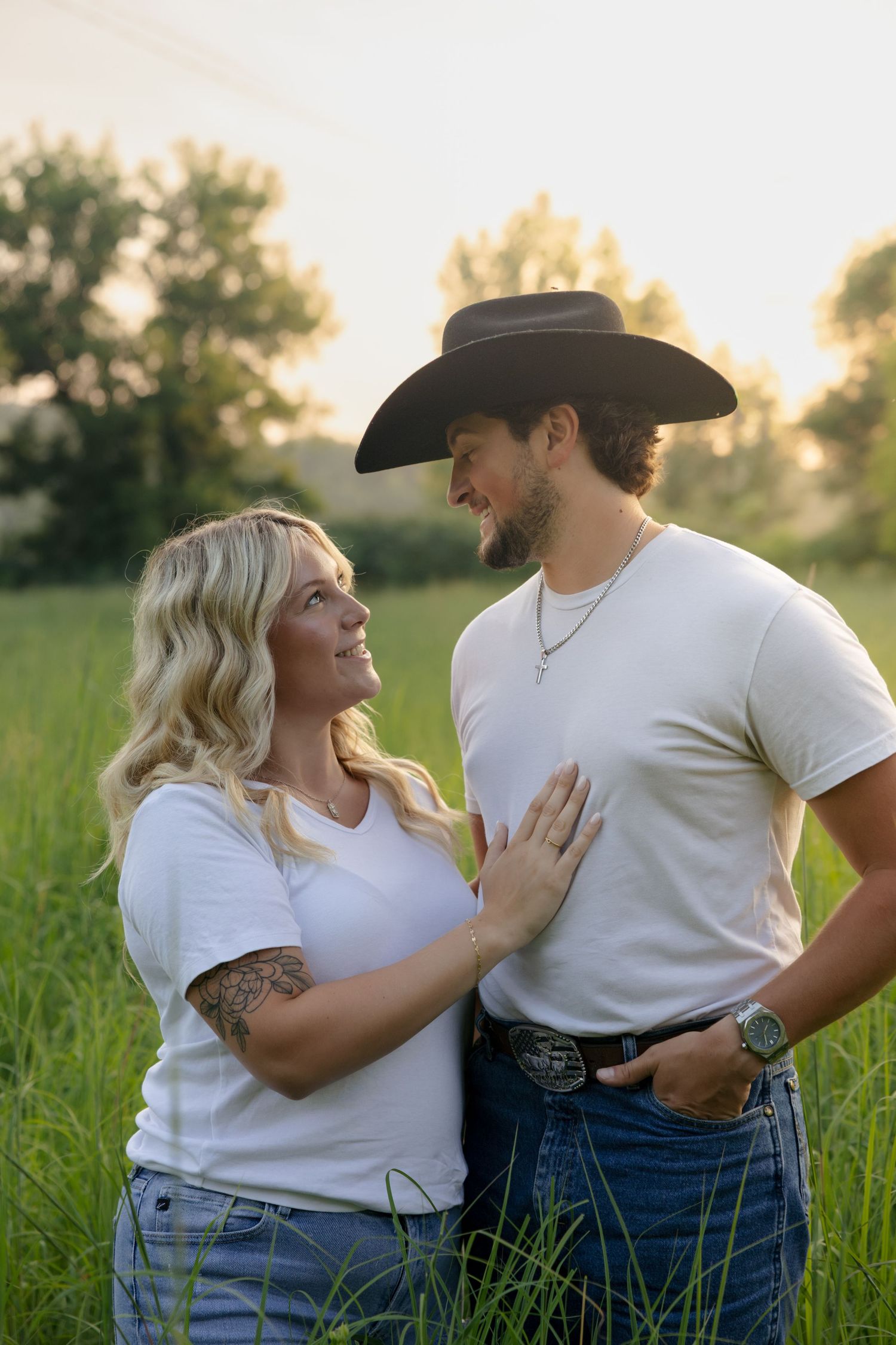 A couple in white tees and jeans share a romantic moment in a sunlit green field, man wearing a black cowboy hat.