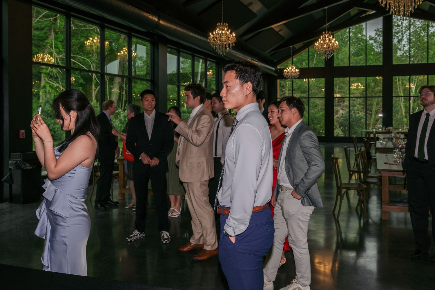 Guests in formal attire gather inside an elegant glass-walled venue surrounded by lush greenery during a wedding reception.