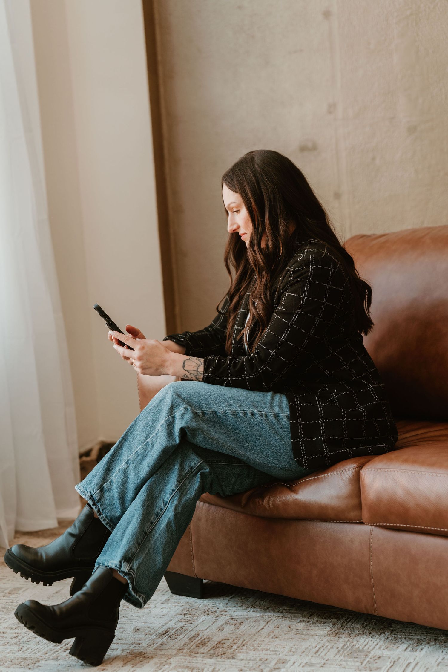 A young woman in jeans and a plaid blazer sits on a brown leather sofa, smiling while using her smartphone.