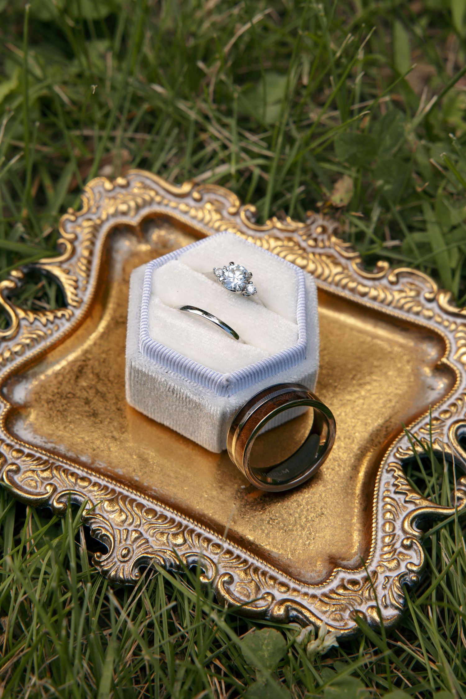 Diamond engagement ring and wedding bands displayed on a velvet box atop a gold ornate tray in green grass.