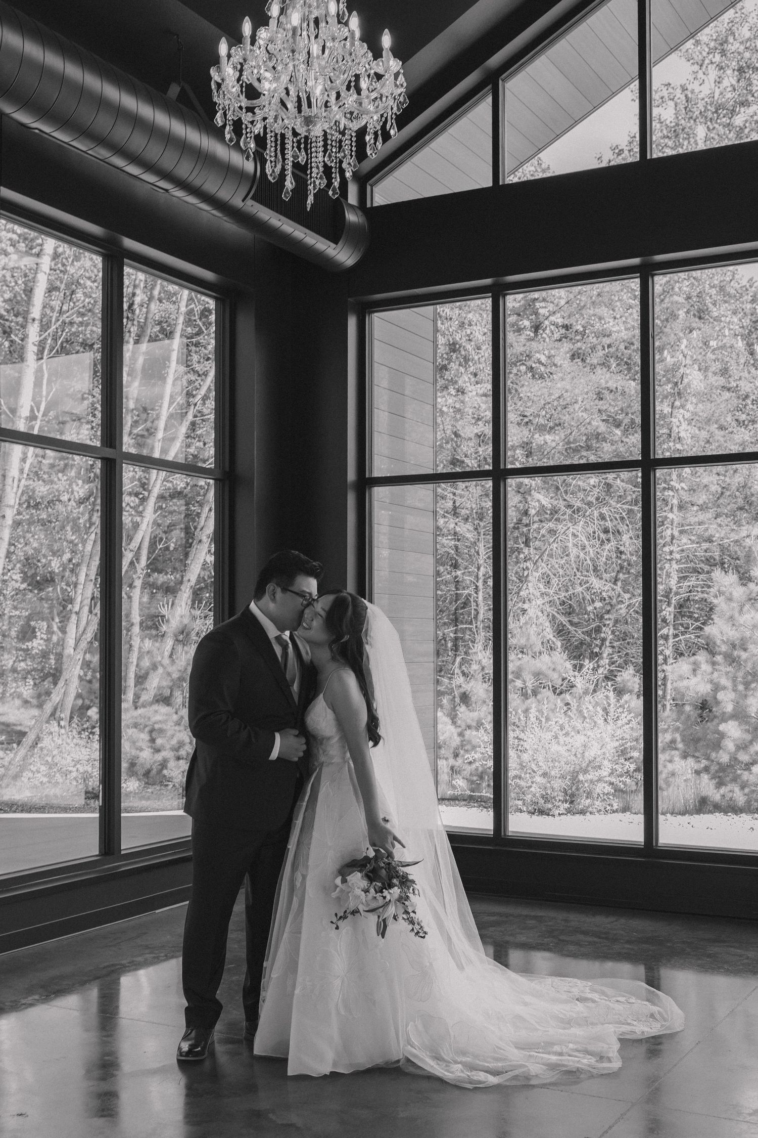 Black and white photo of a bride and groom sharing a kiss under a chandelier by large windows in an elegant venue.