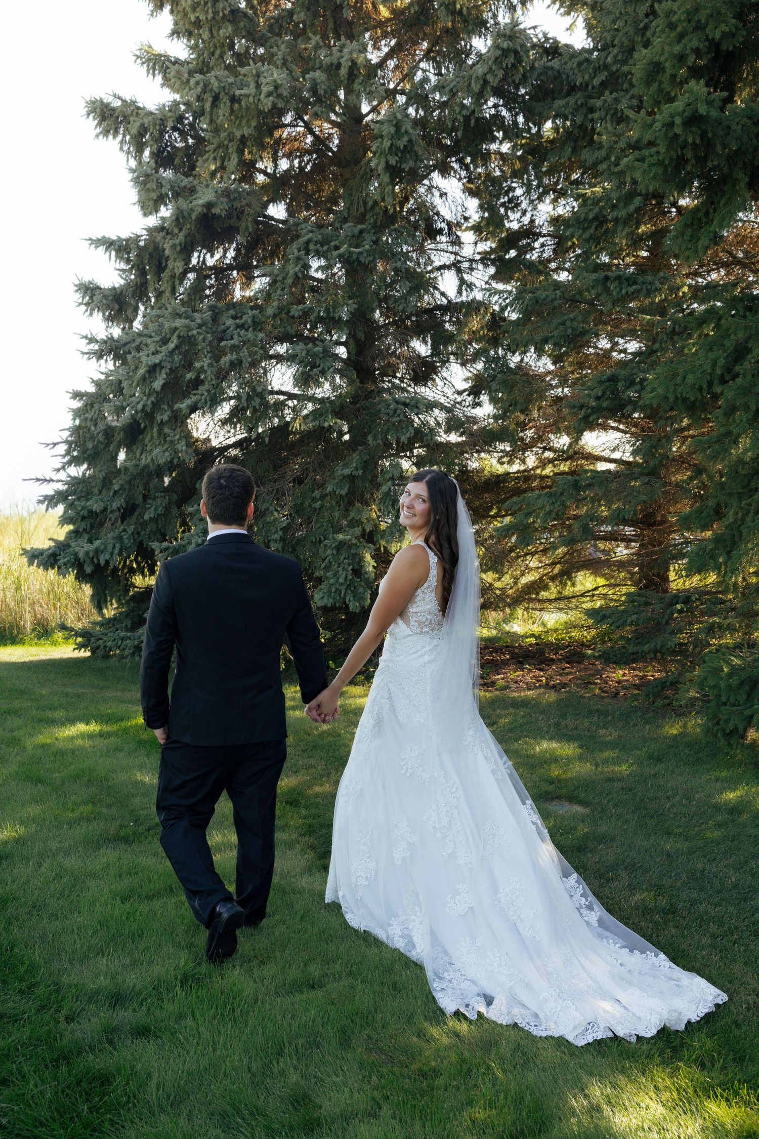 Bride in lace gown holding grooms hand walking through green lawn near tall pine trees.