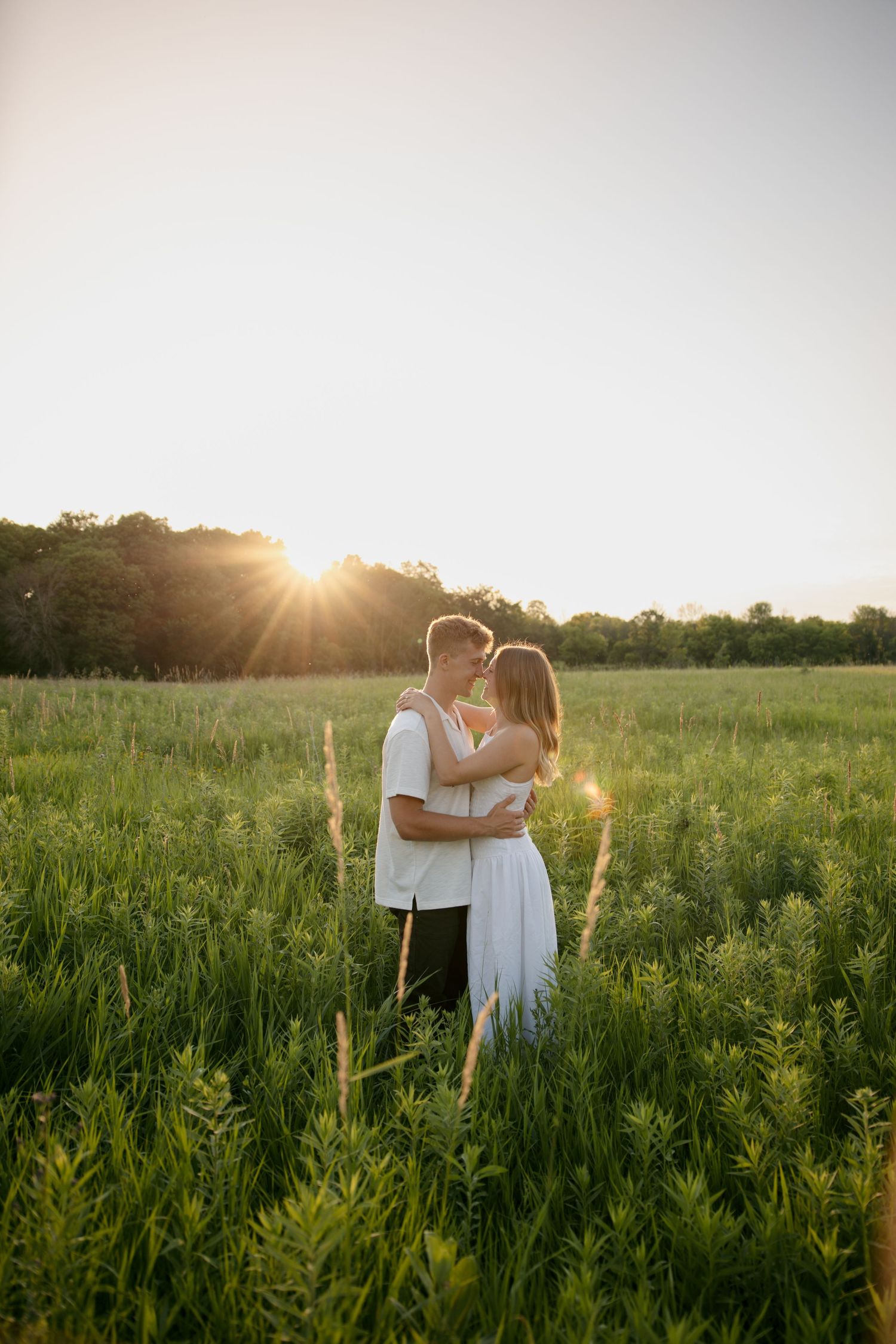 Romantic couple embracing in a green field at golden hour sunset with warm sun rays glowing behind them.