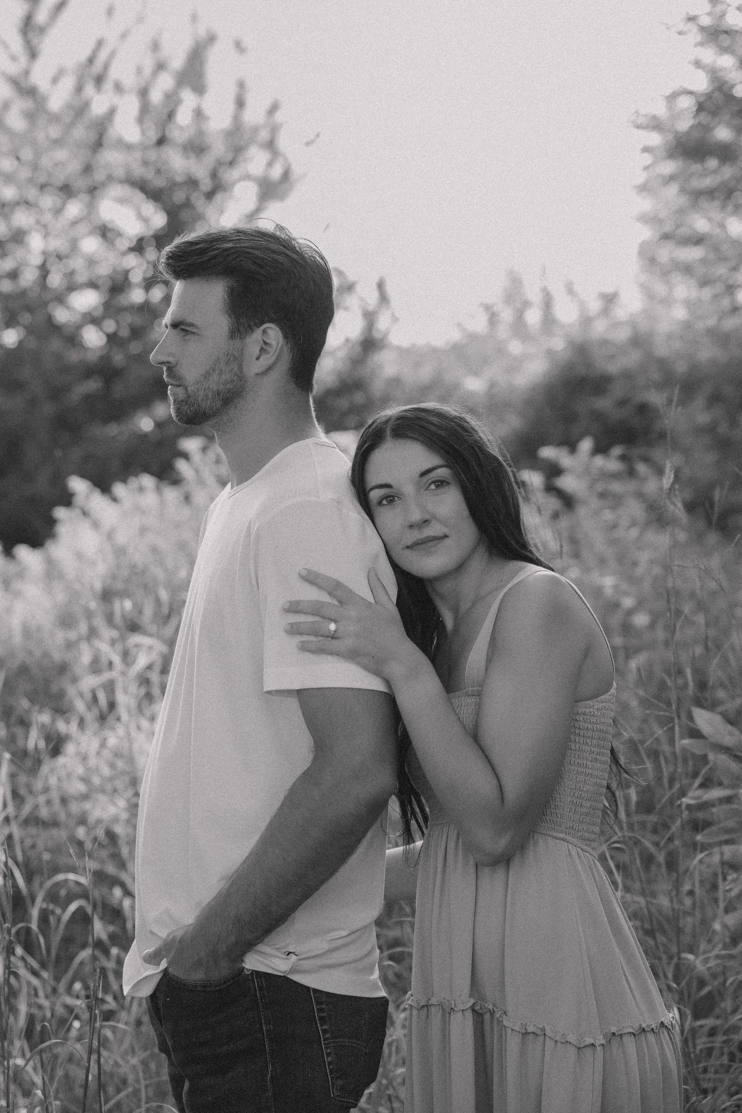 Black and white photo of a couple embracing outdoors, woman in dress leaning on mans shoulder among lush greenery.