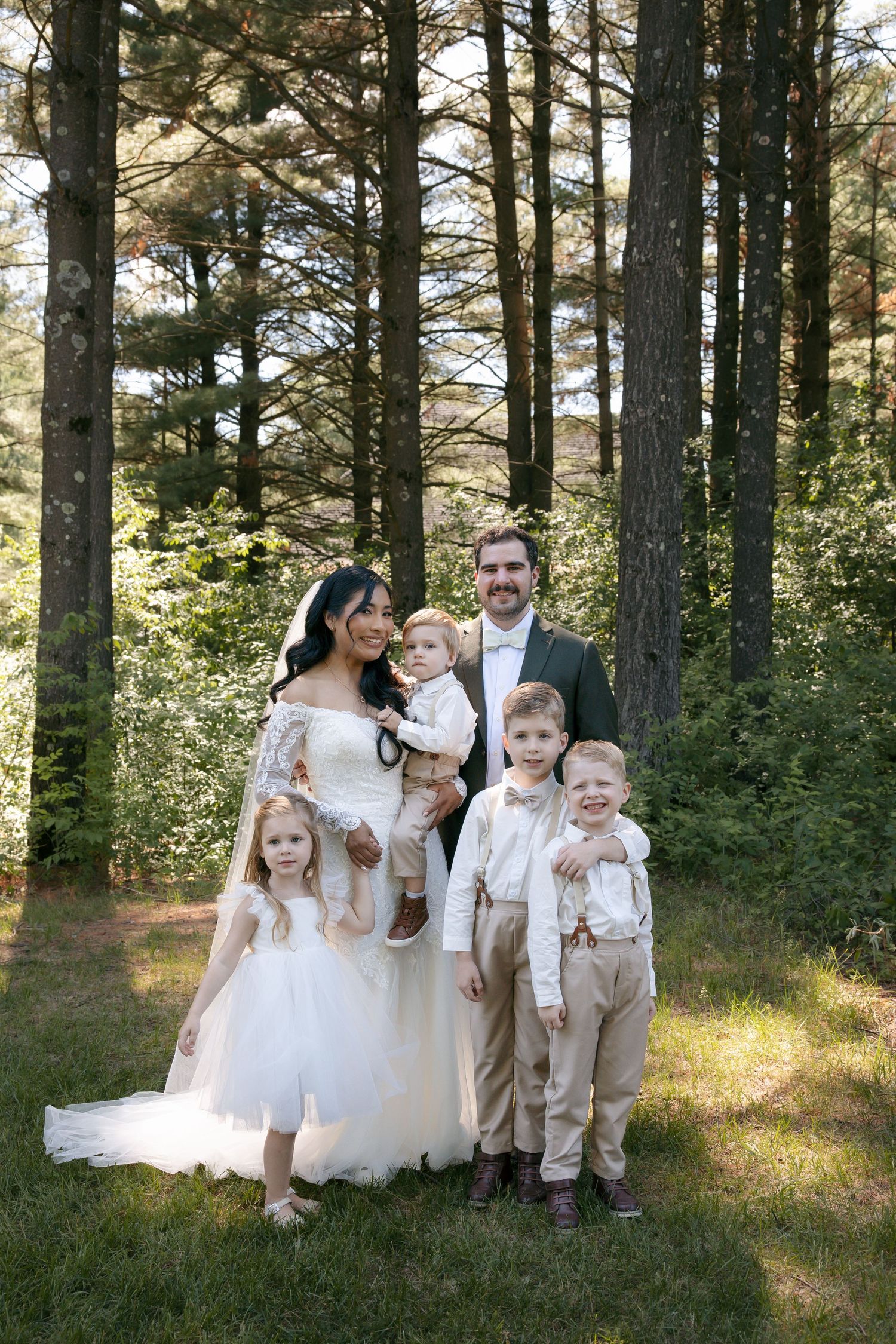 Bride and groom pose with three young children in wedding attire among tall pine trees on their wedding day.