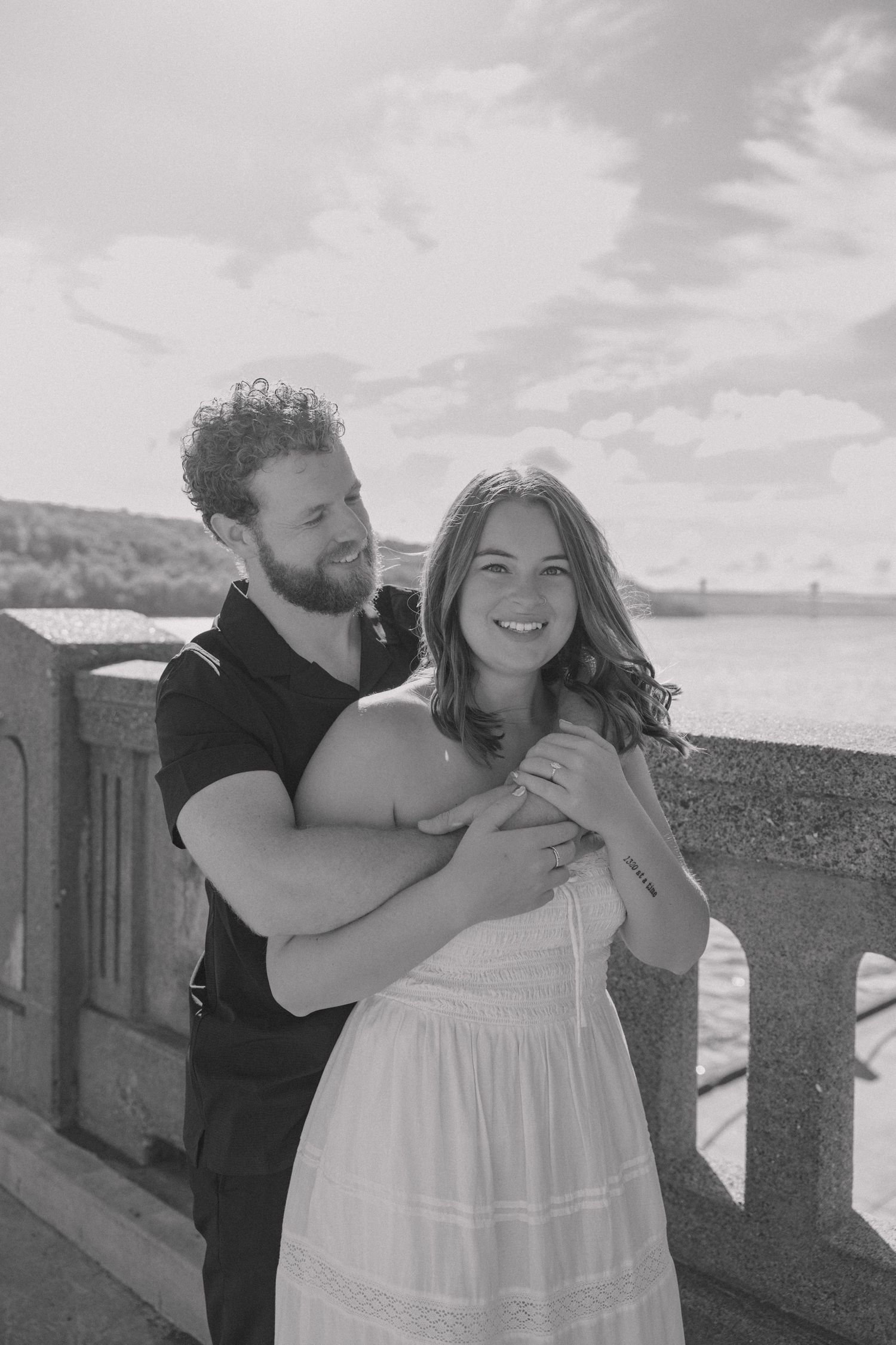 Black and white photo of a smiling couple embracing on a bridge, woman in white dress, man in black shirt.