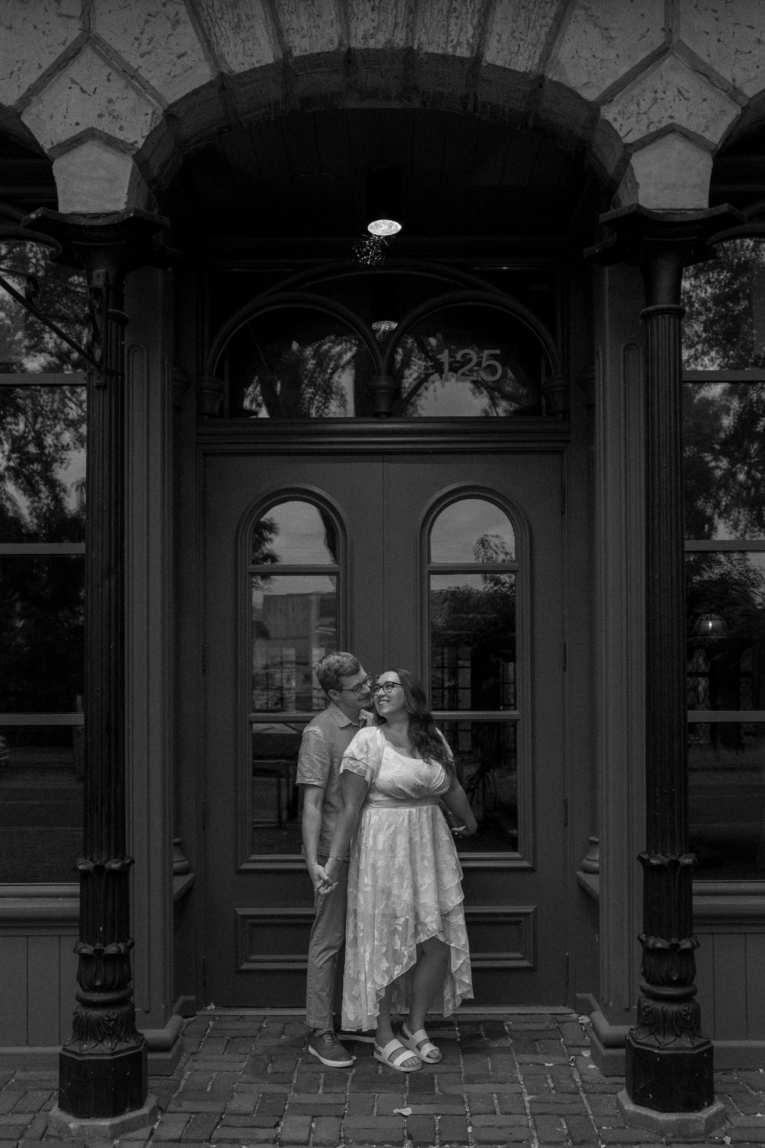 Black and white photo of a couple holding hands and smiling in front of an elegant arched doorway.
