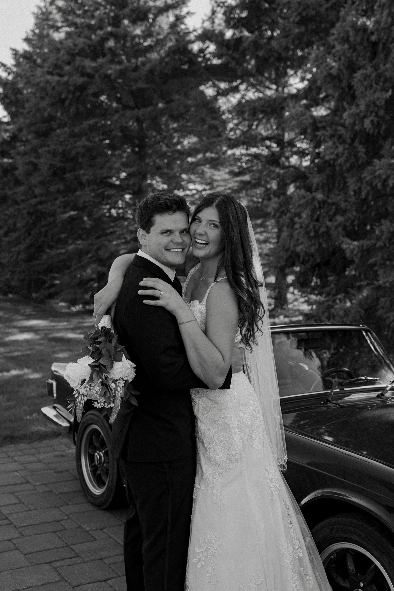 Black and white portrait of smiling bride and groom embracing beside a classic vintage car outdoors.